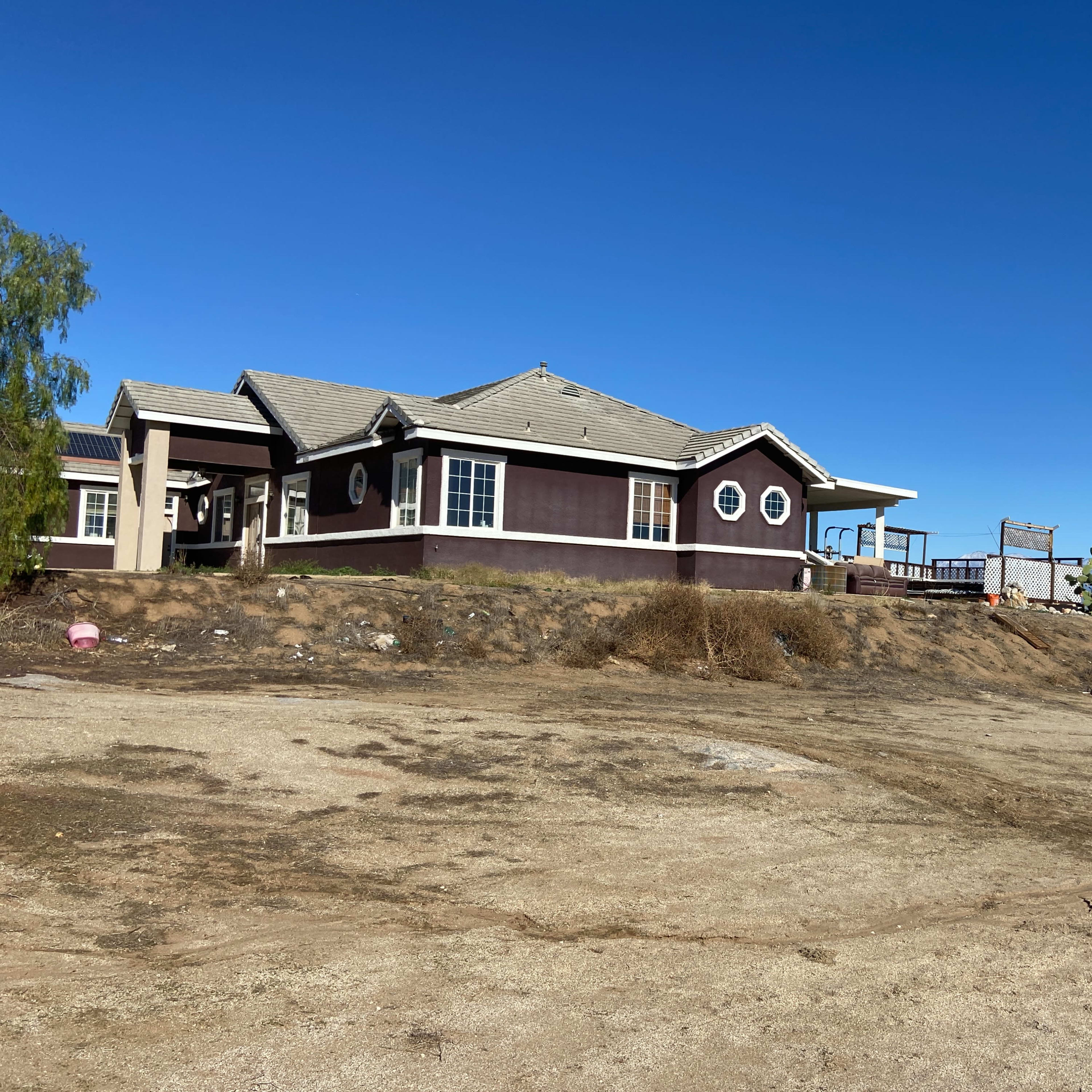 A large house with a brown exterior and multiple windows sits on a barren lot under a clear blue sky.