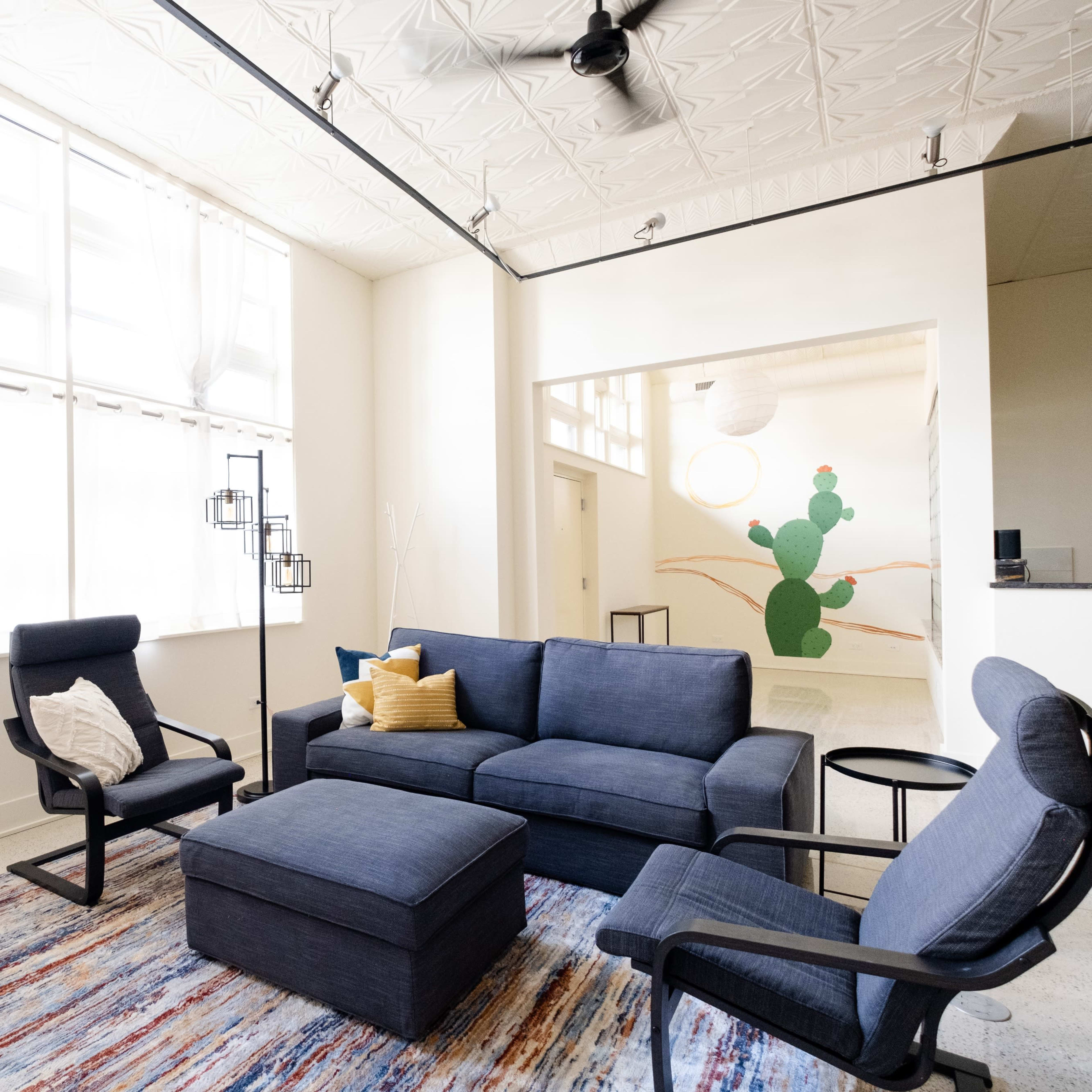 A modern living room features a navy blue sofa and chairs, a patterned rug, large windows with light curtains, and a green cactus mural in the background.