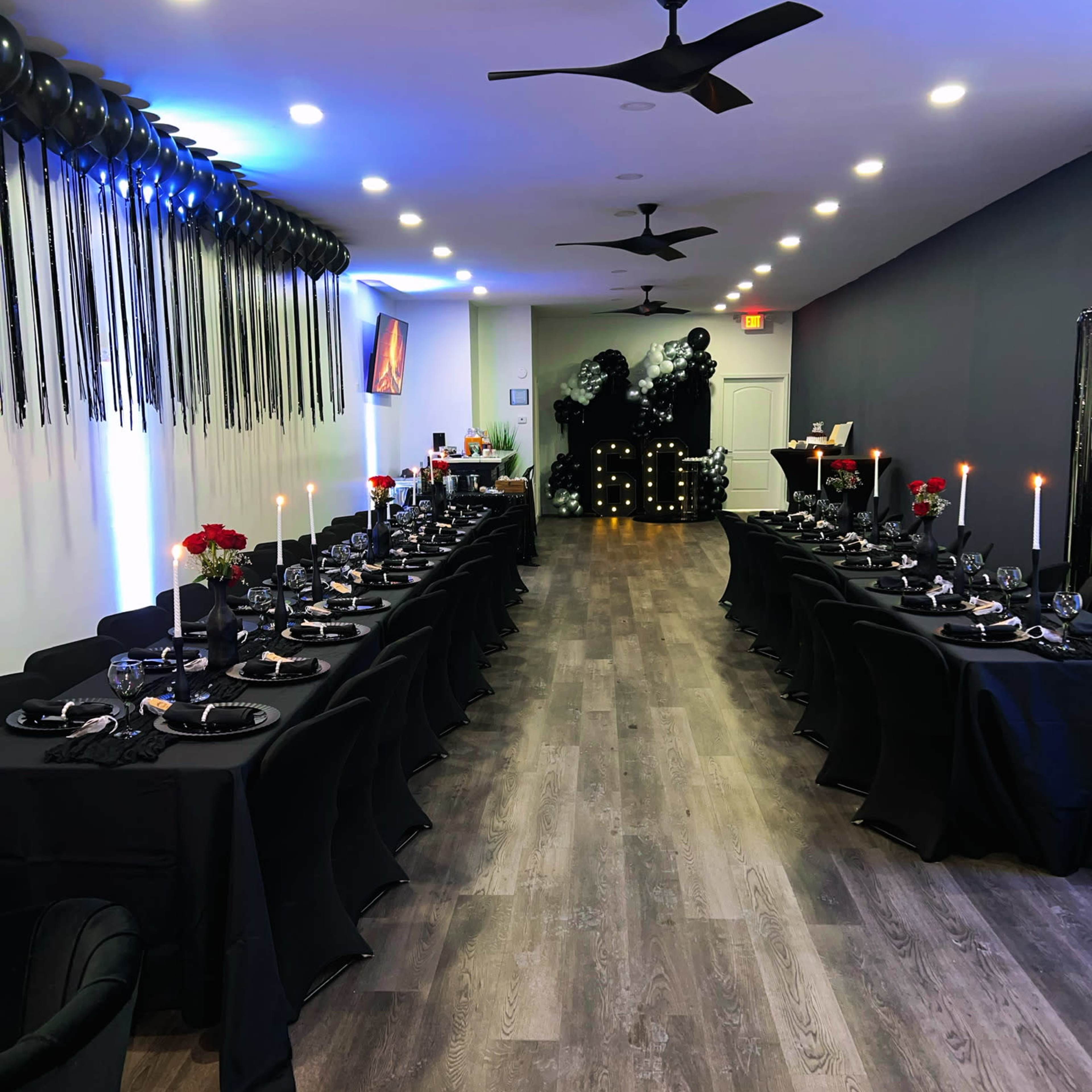 A long, elegantly set dining table is arranged in a dimly lit room with black tablecloths, candles, and decorative balloons along the walls.