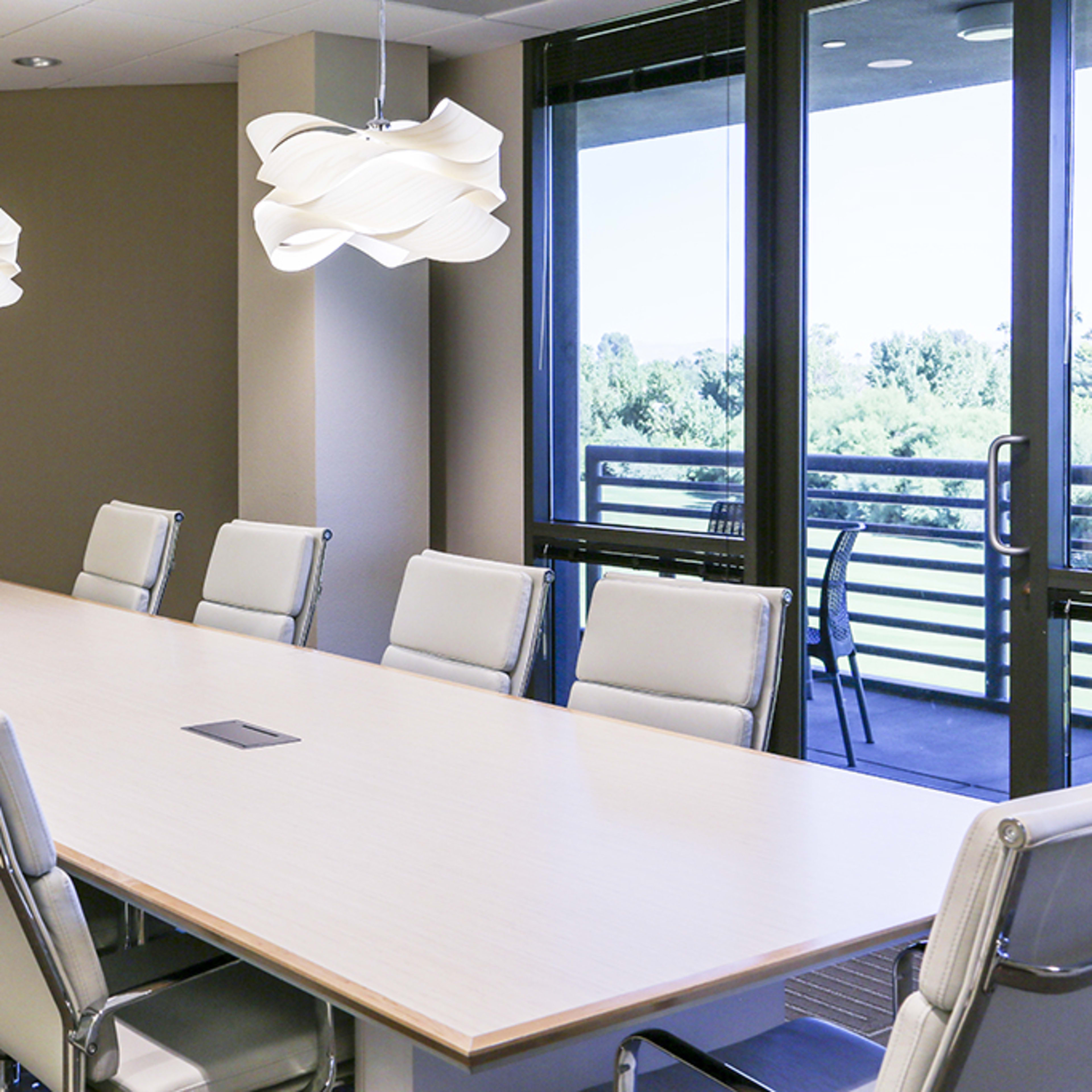A modern conference room features a long, light-colored table surrounded by leather chairs and illuminated by designer light fixtures.
