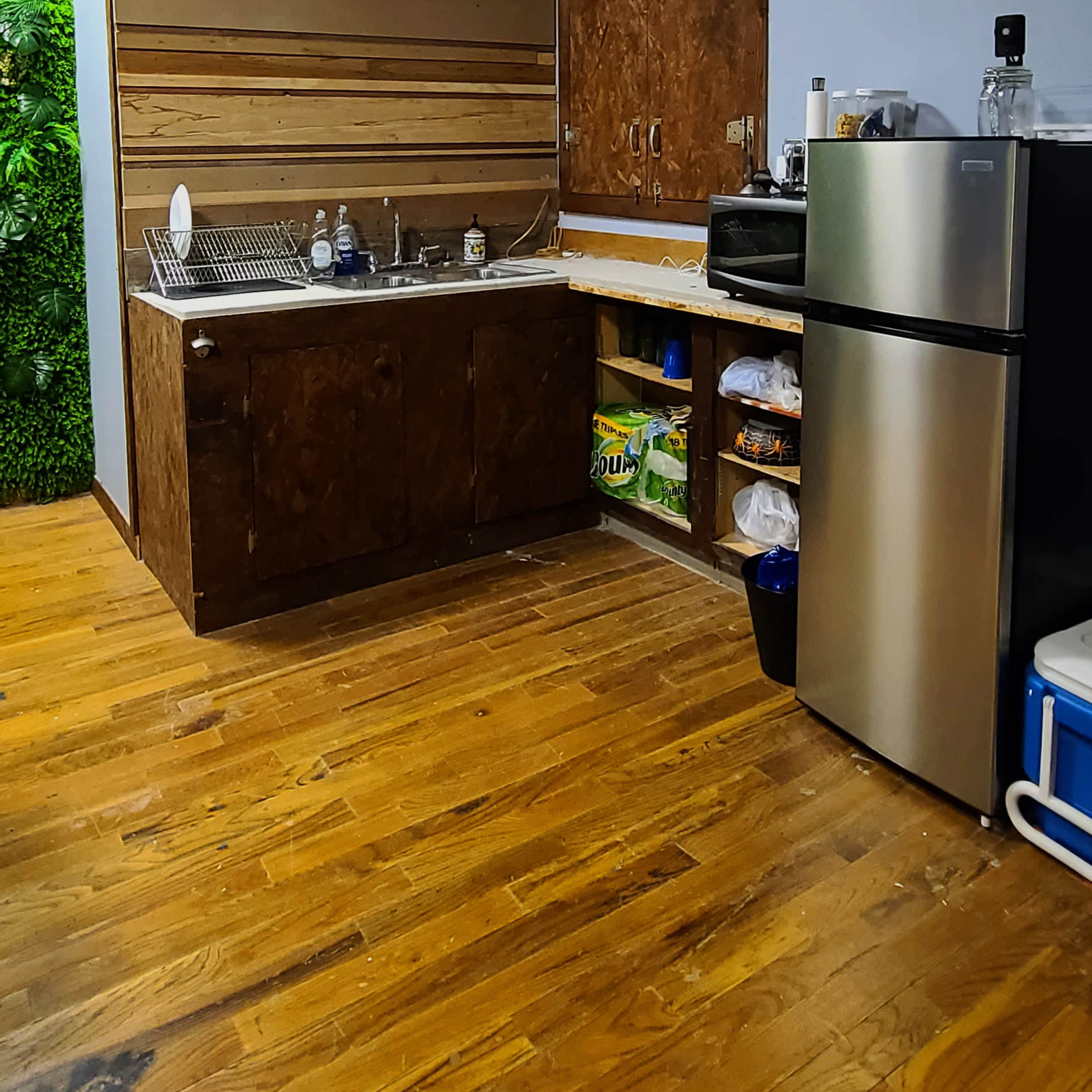 A kitchen area with a sink, wooden cabinets, a refrigerator, and a few shelves against a wall, featuring wooden flooring and a partially visible green wall.