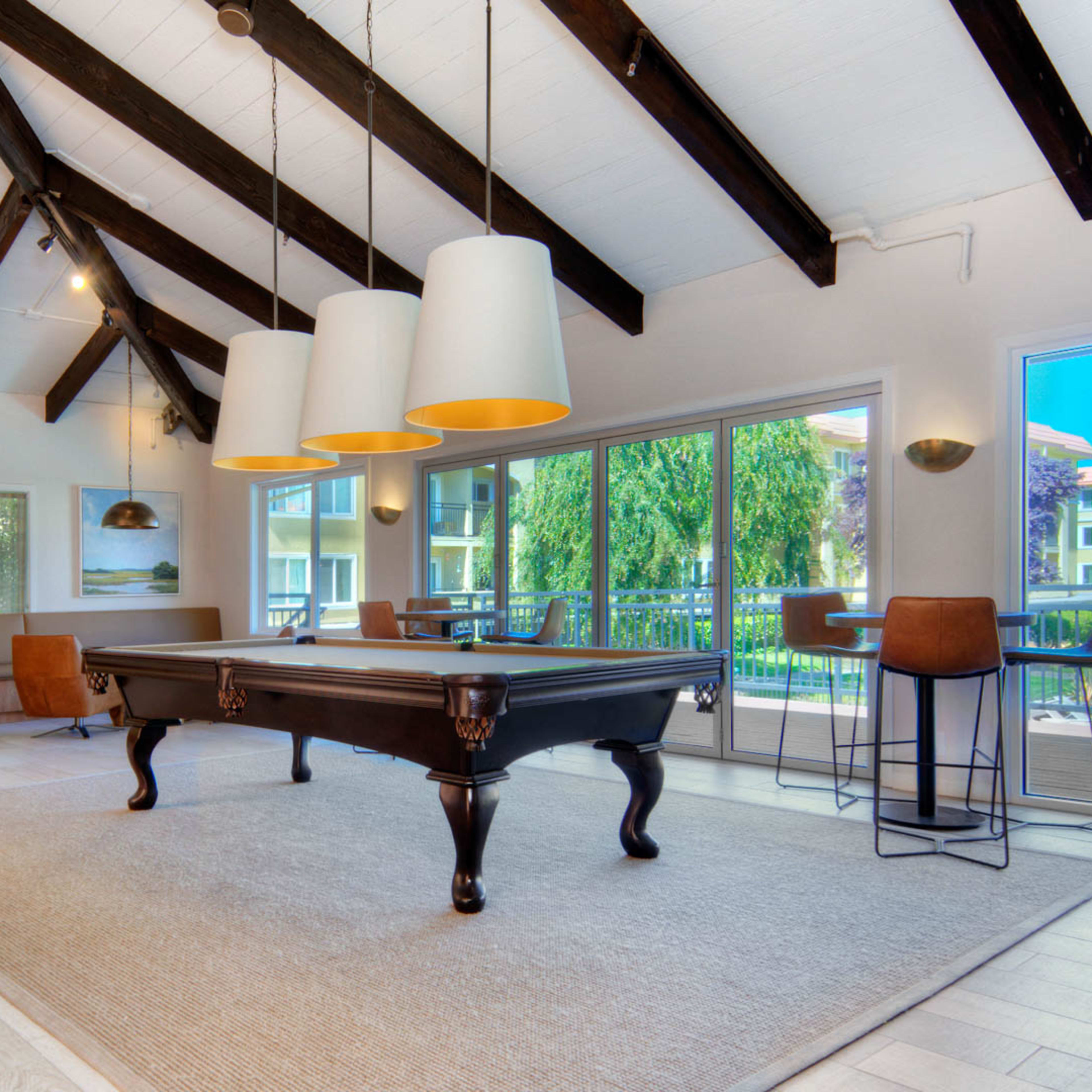 A spacious room features high vaulted ceilings, a billiard table, and large windows overlooking an outdoor area.