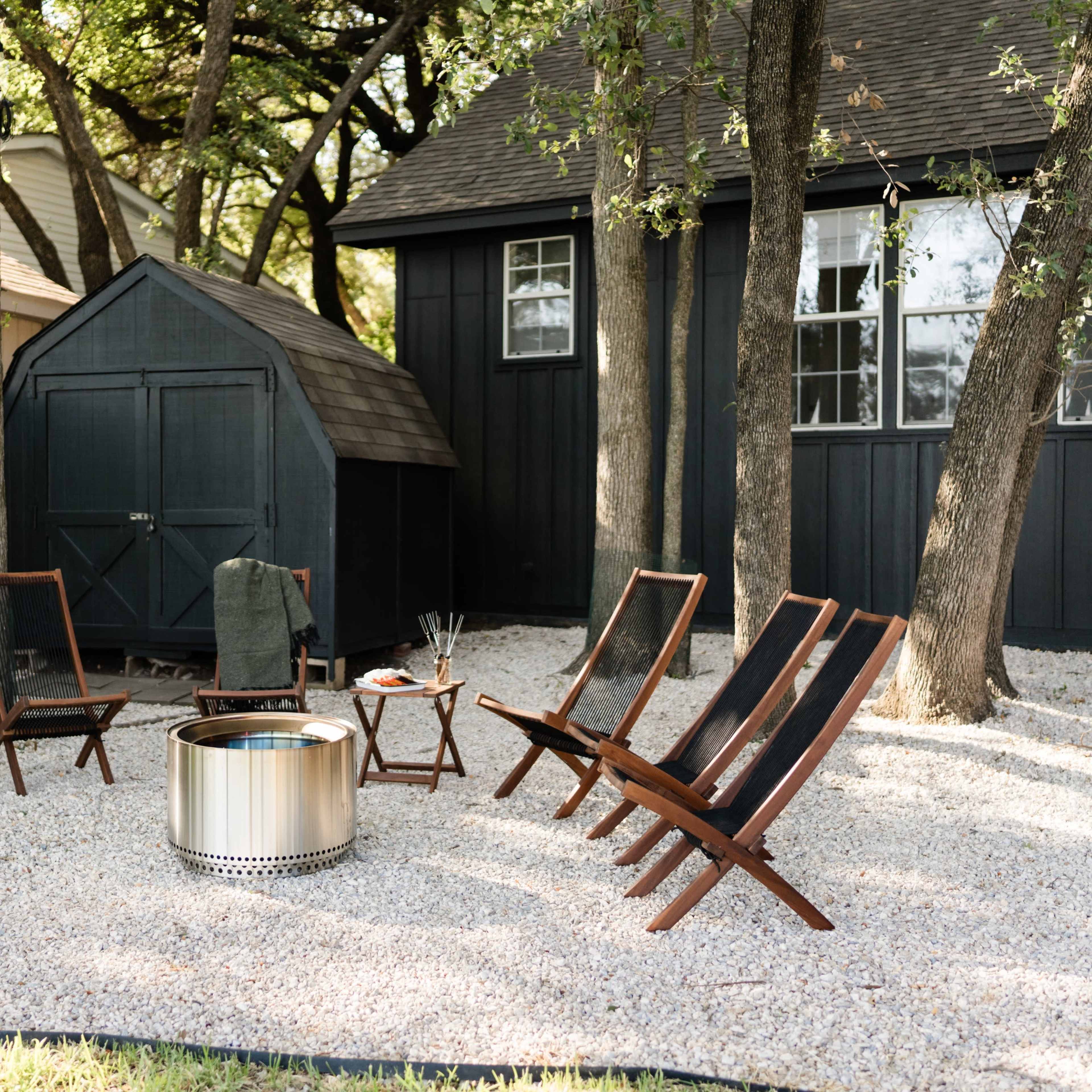 A circular fire pit surrounded by wooden chairs and a small table is situated on a gravel patio next to a shed and kayaks in a backyard.