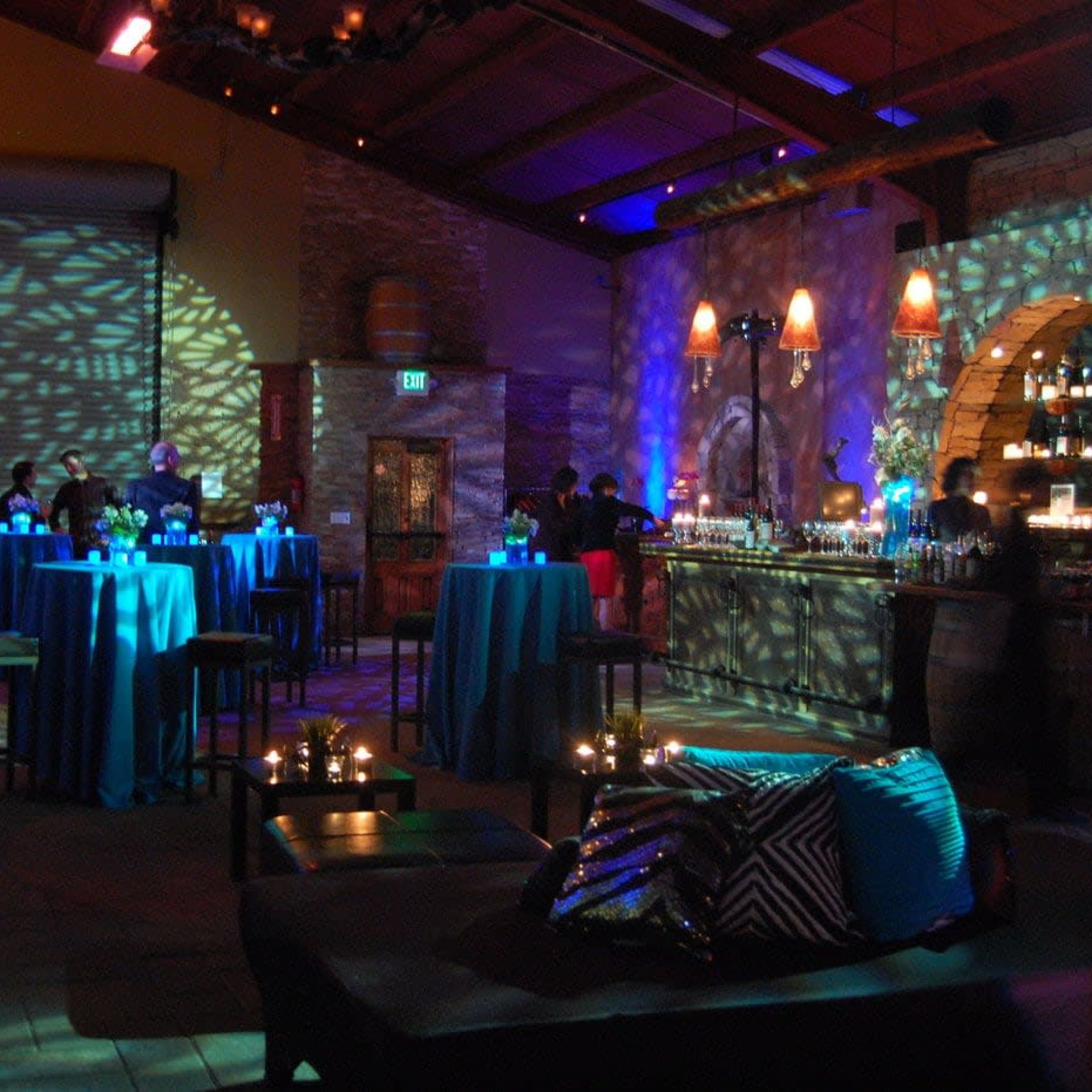 A dimly lit event space with high tables draped in blue cloth, a bar area in the background, and decorative lighting creating patterns on the walls.
