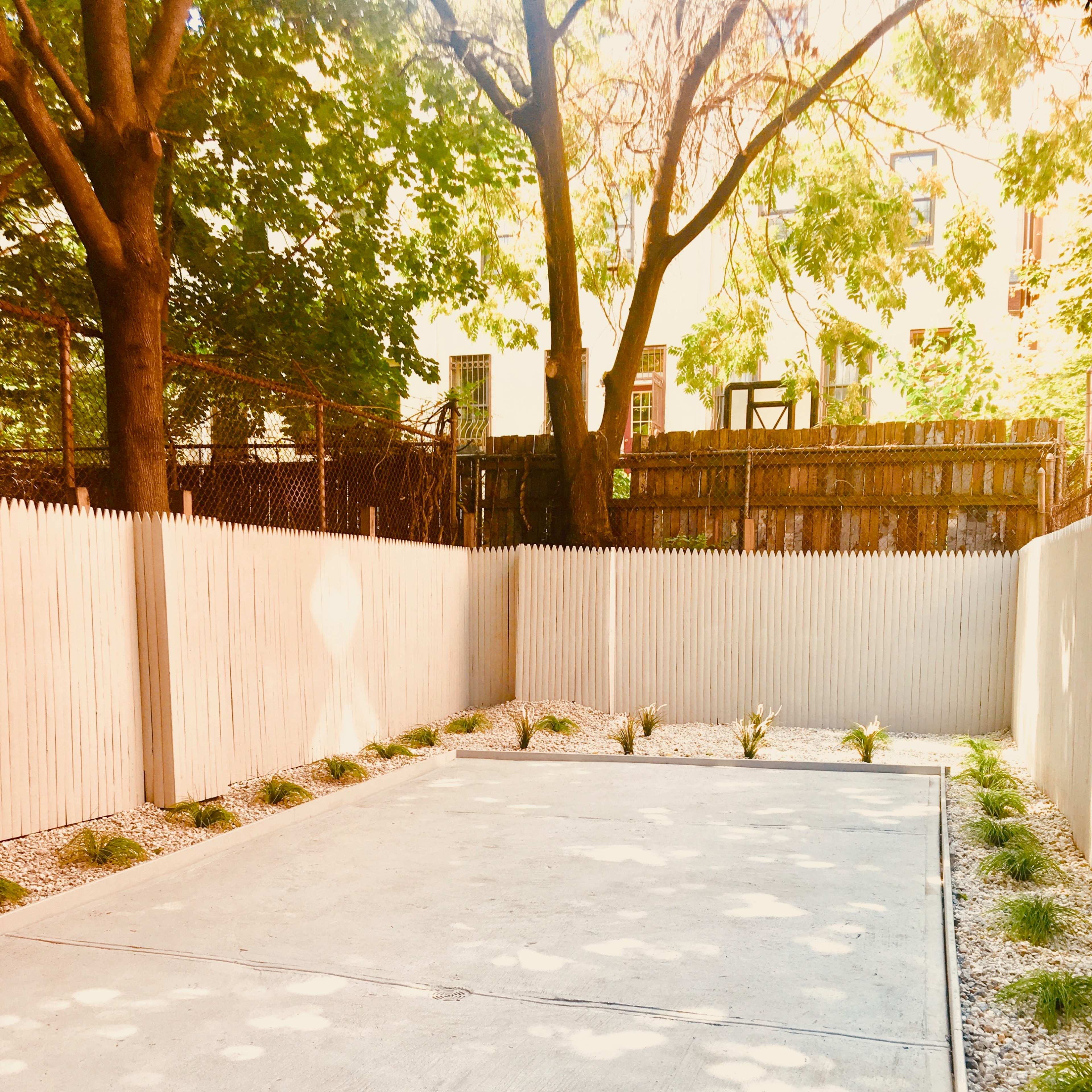 A concrete outdoor space is bordered by a white picket fence and features small plants along the edges.