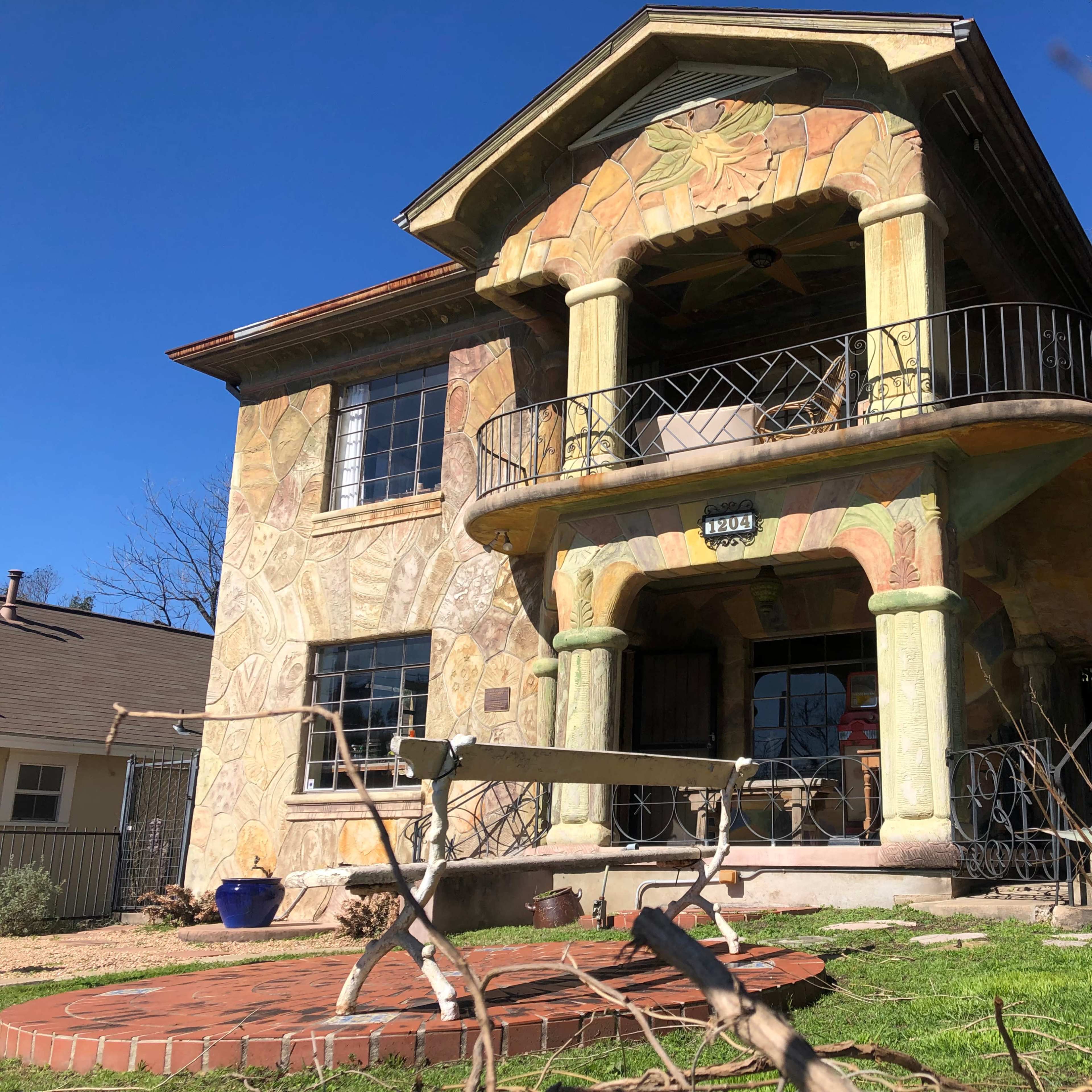 A two-story house with colorful mural-like decorations on its exterior, featuring a balcony supported by decorative columns and surrounded by a garden.