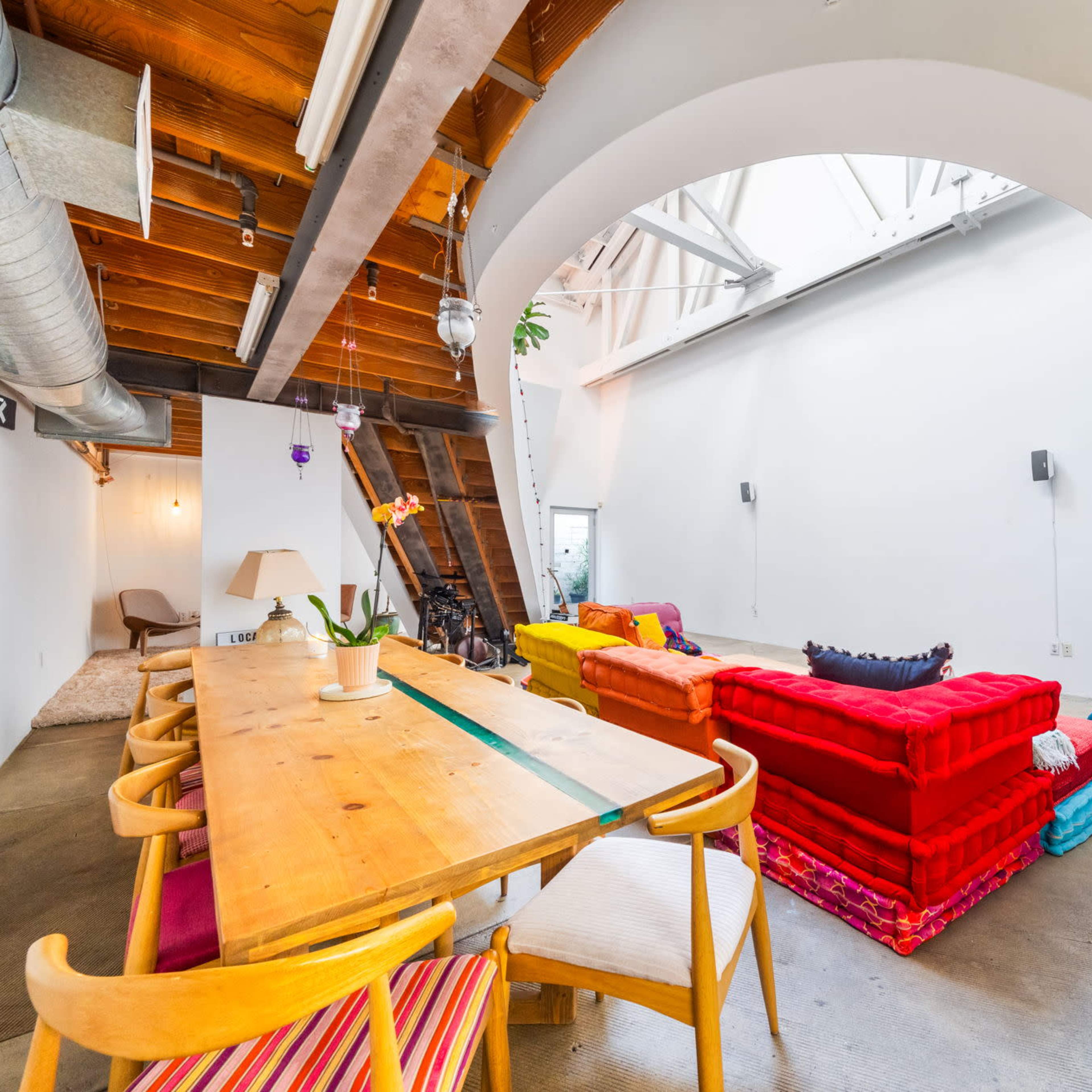 A spacious living area with a wooden dining table, colorful sofas arranged in a semi-enclosed space, and a high ceiling with exposed beams.