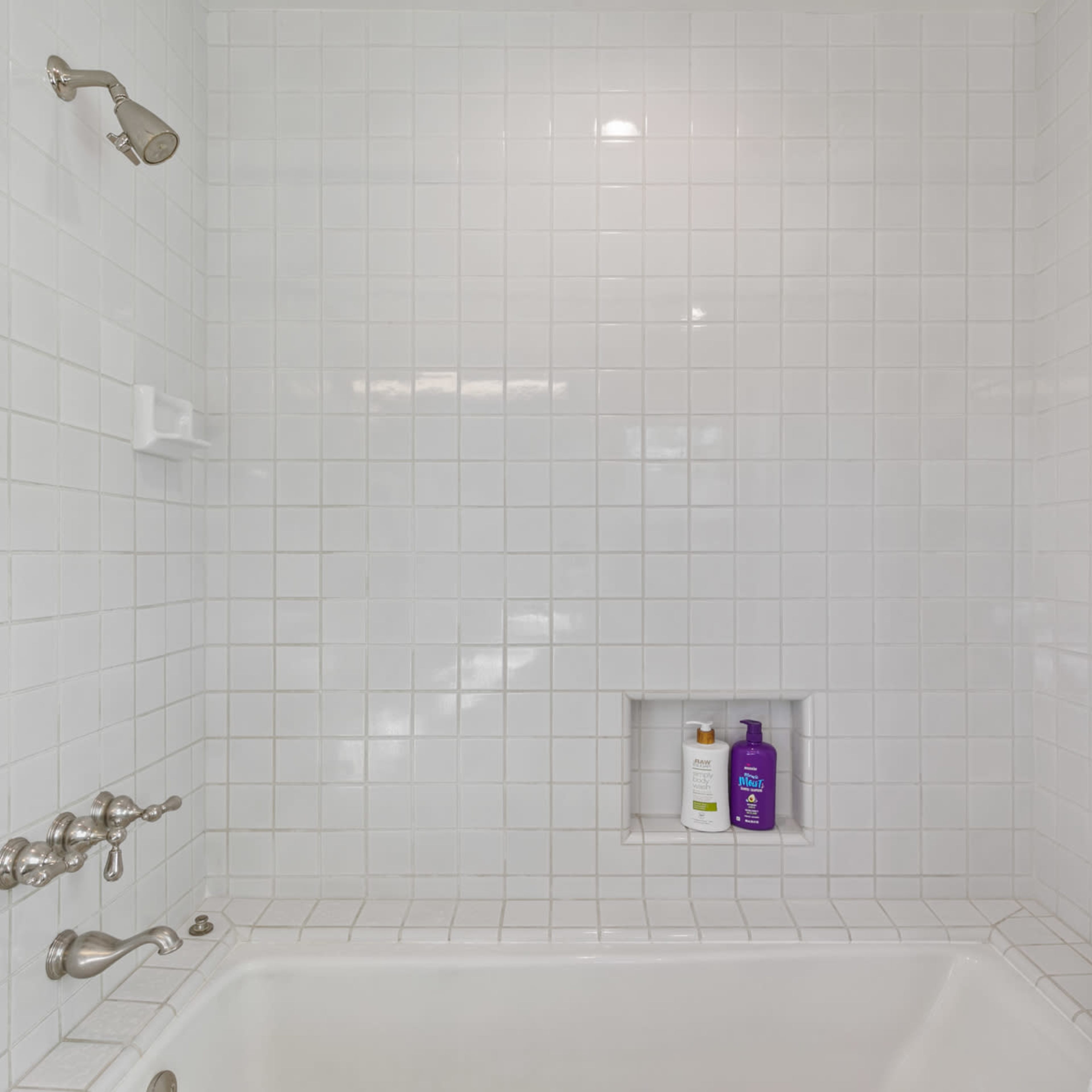 A clean white tiled shower with a showerhead, grab bars, and shampoo bottles neatly arranged in a wall recess.