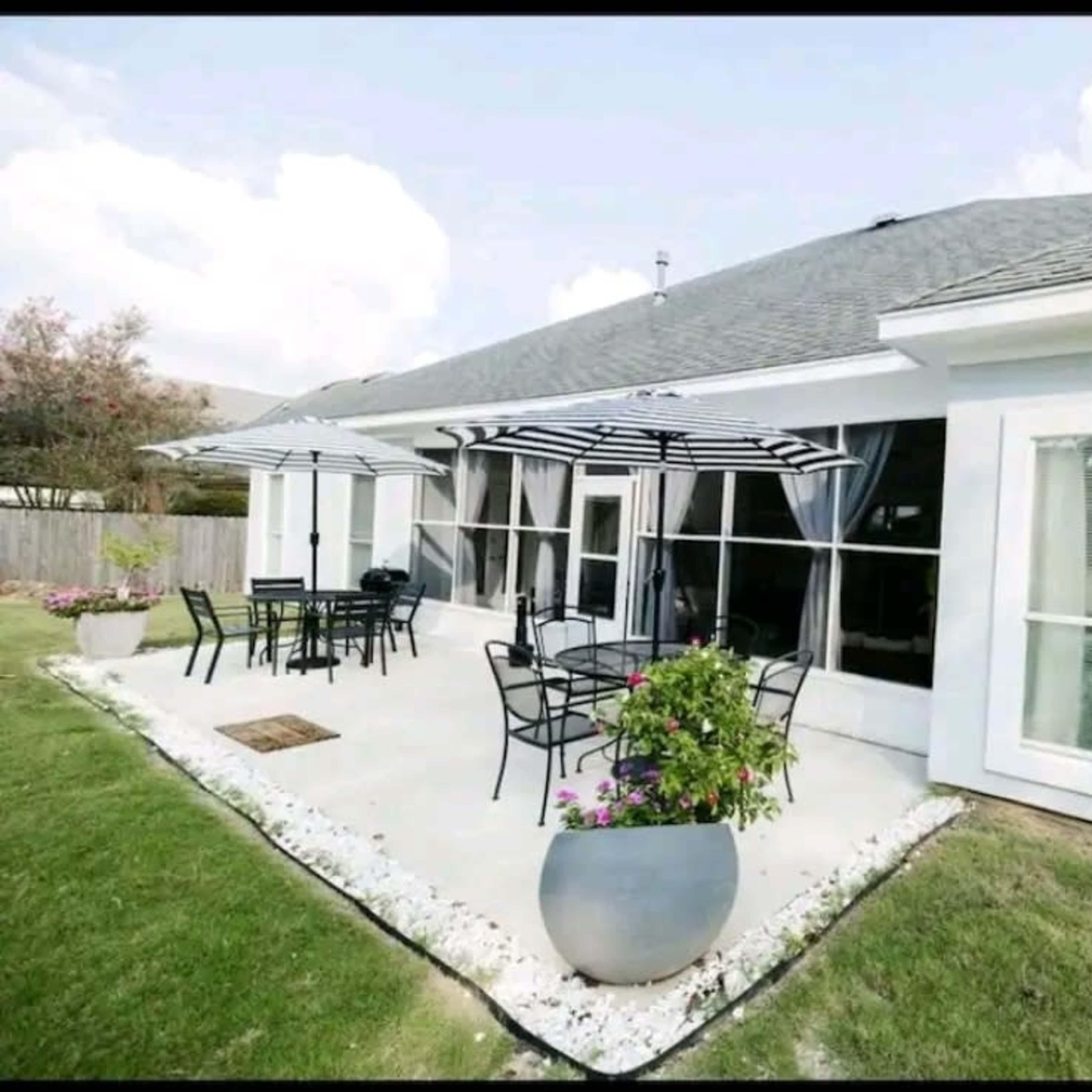 A modern patio area with metal furniture, large umbrellas, and flowerpots, adjacent to a house with large windows.