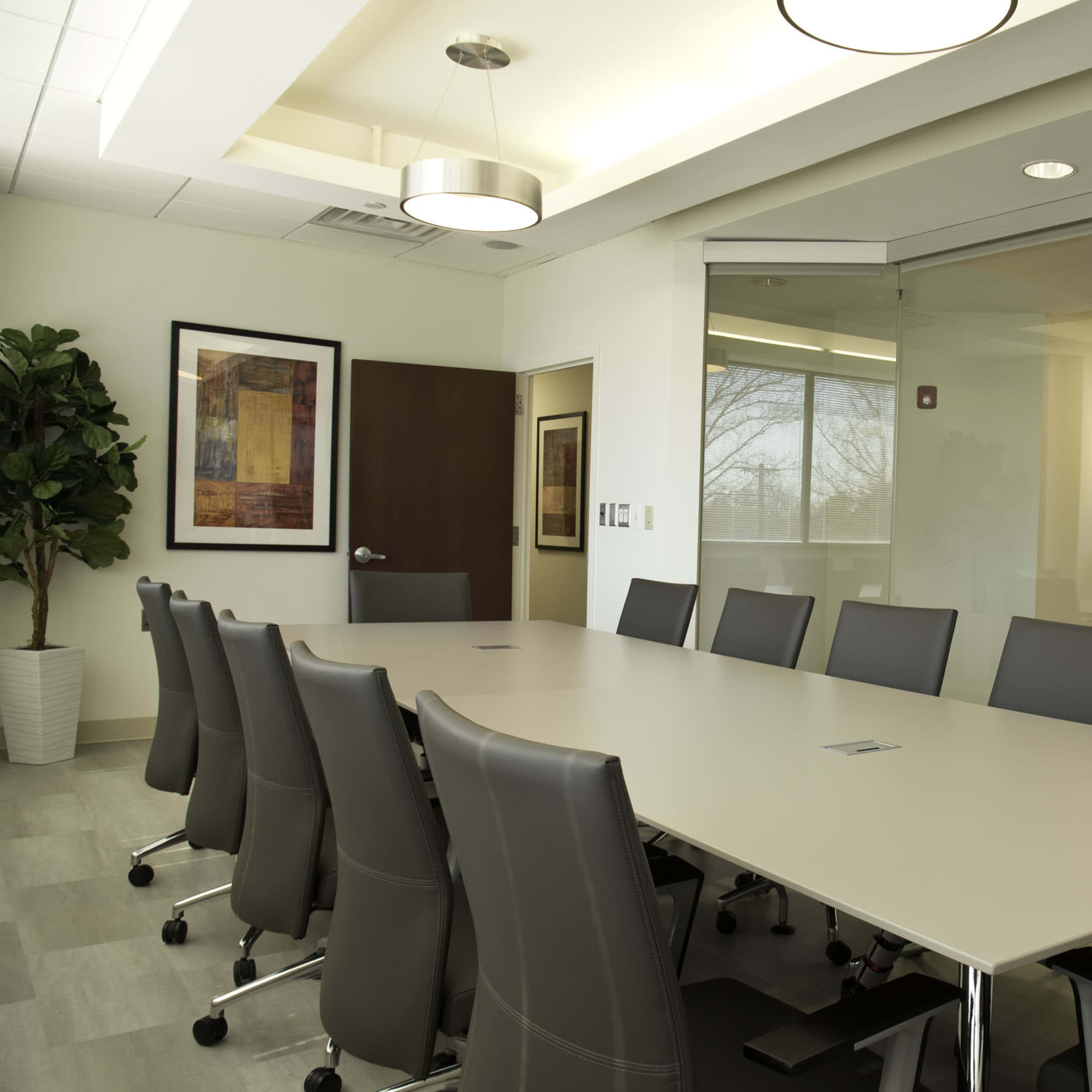 A modern conference room features a large rectangular table surrounded by ergonomic chairs and a glass wall offering a view of an outside landscape.