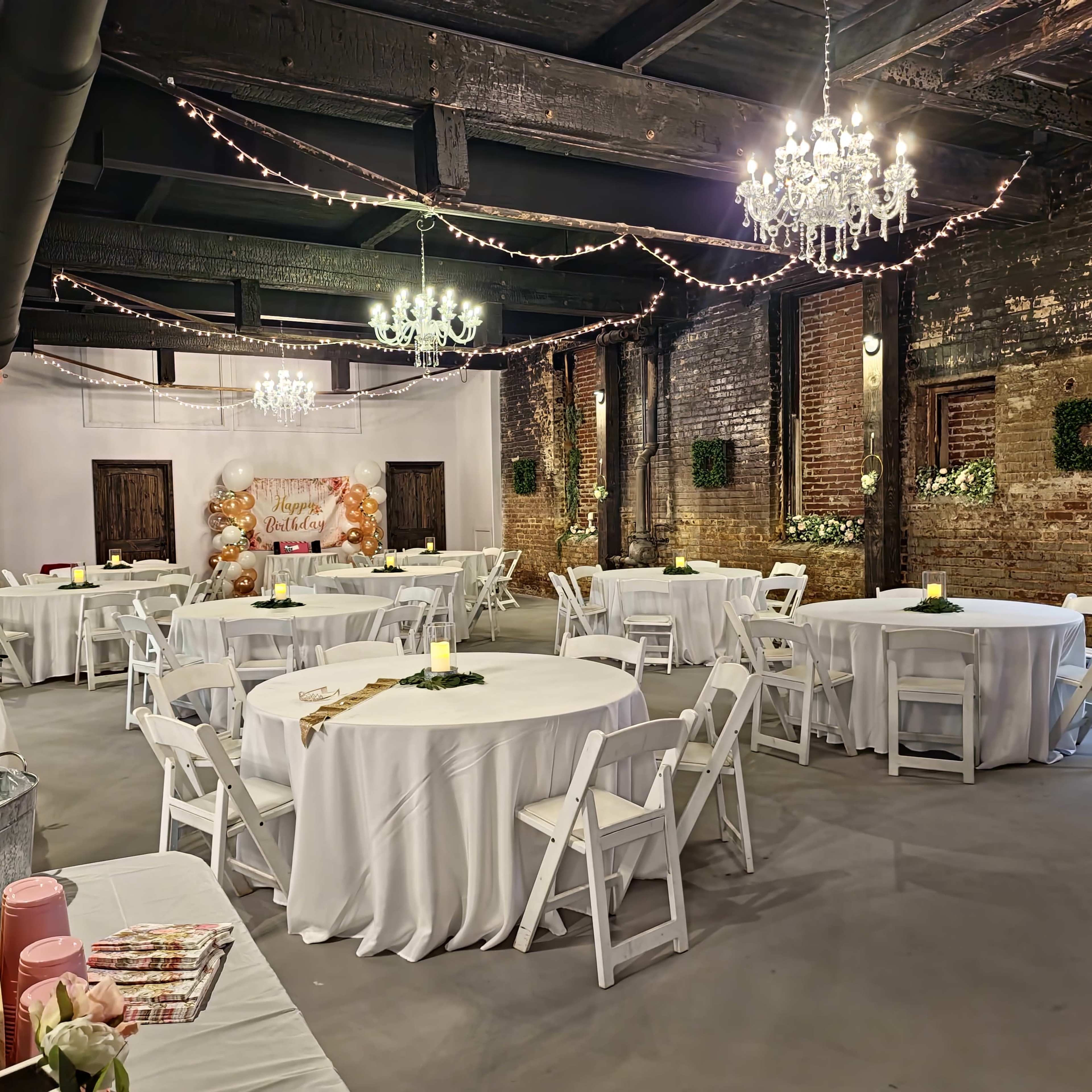 A decorated event space features round tables with white tablecloths, string lights overhead, and chandeliers, set against exposed brick walls.