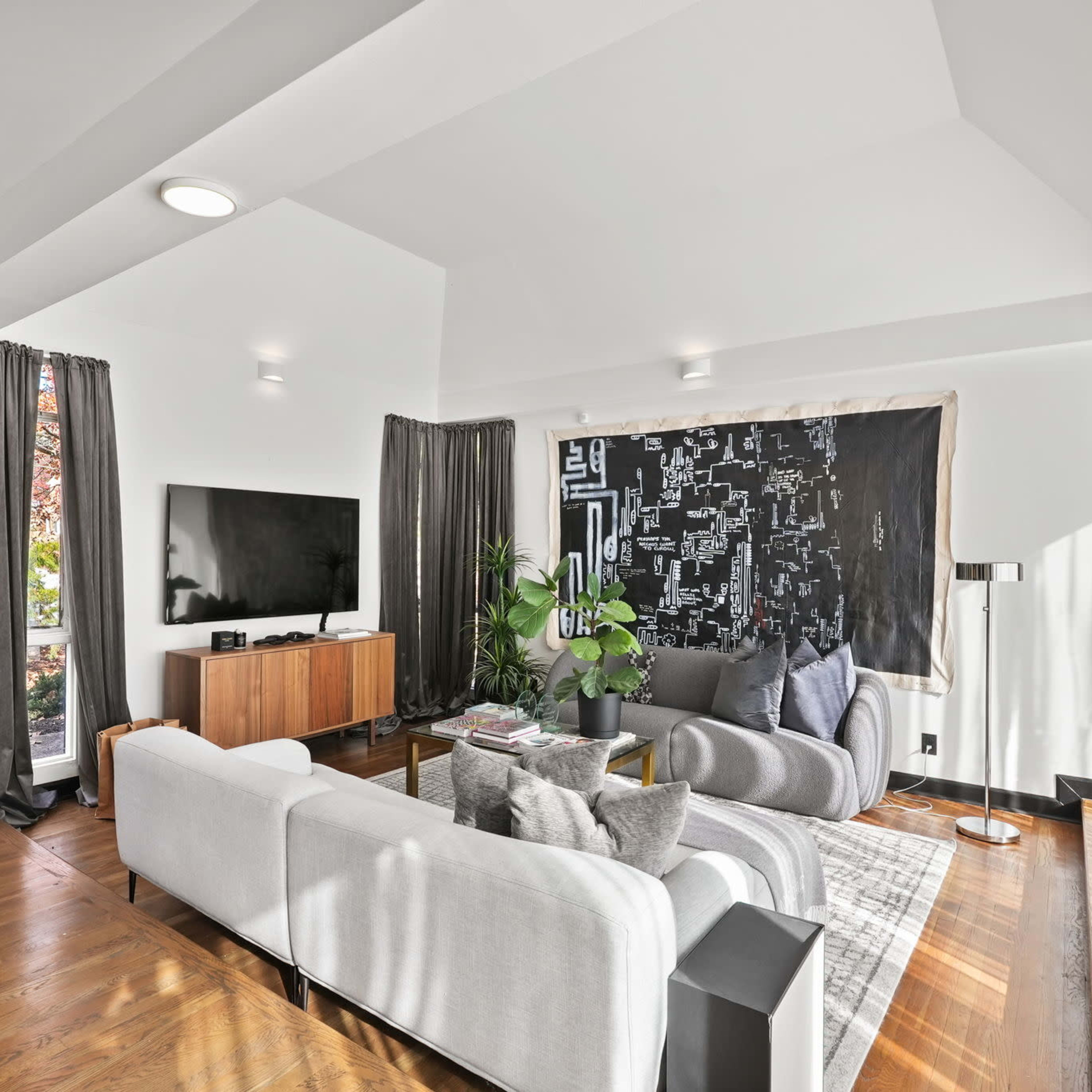 A modern living room featuring a light gray sectional sofa, a black wall art piece, a large television, and a wooden cabinet, with natural light streaming through the windows.