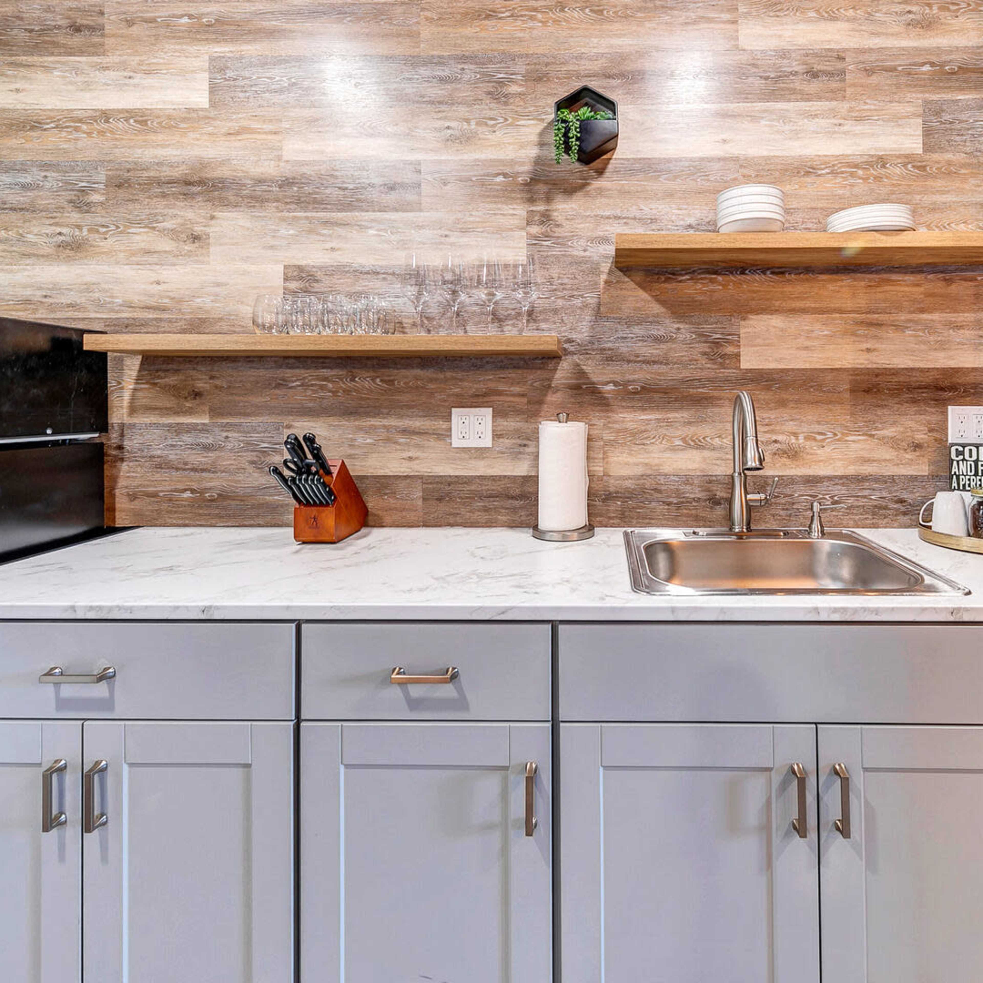 A modern kitchen with wood-panelled walls, a marble countertop, and open shelving displaying dishes and kitchen tools.