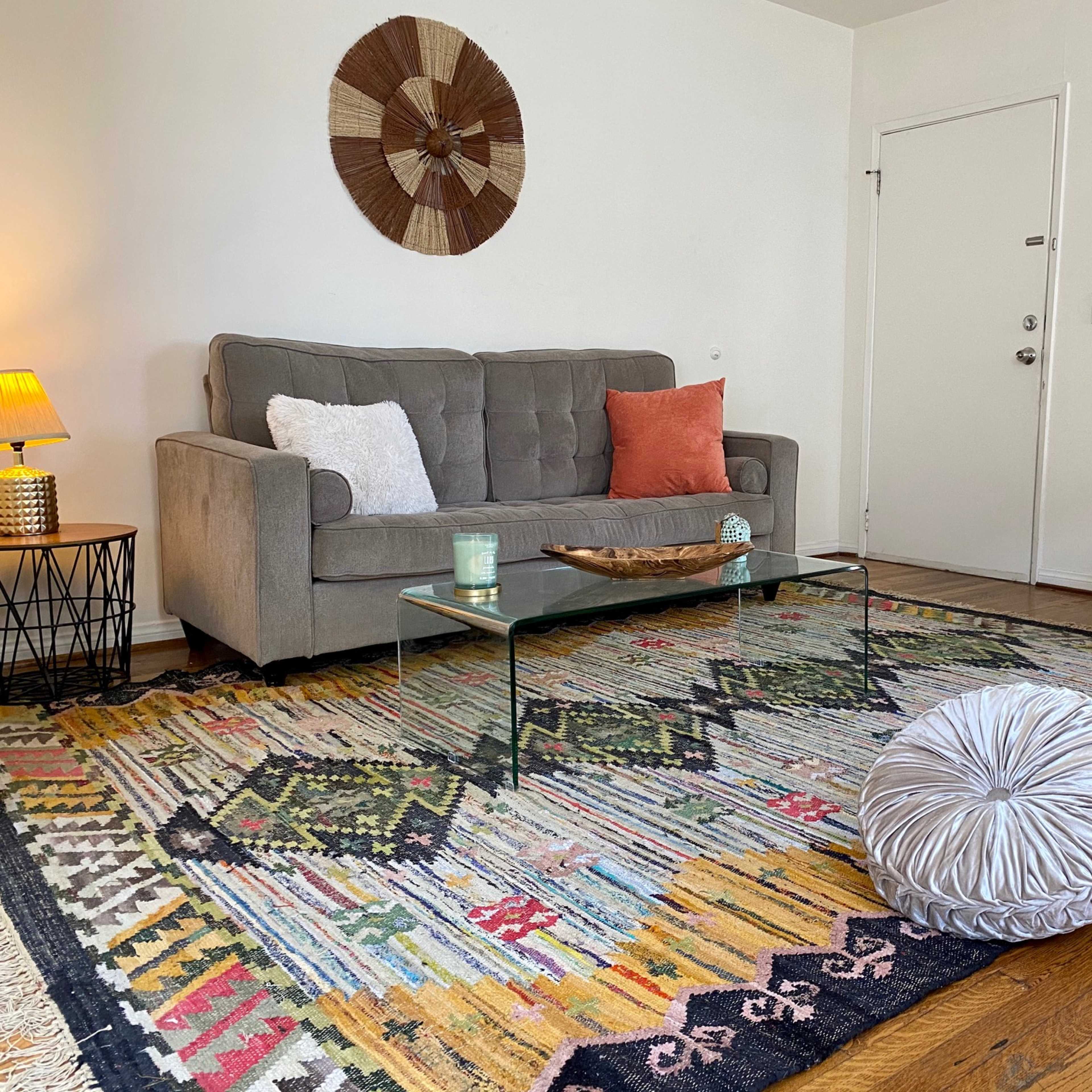 A softly lit living room featuring a gray couch, a glass coffee table, colorful poufs, and a patterned area rug.