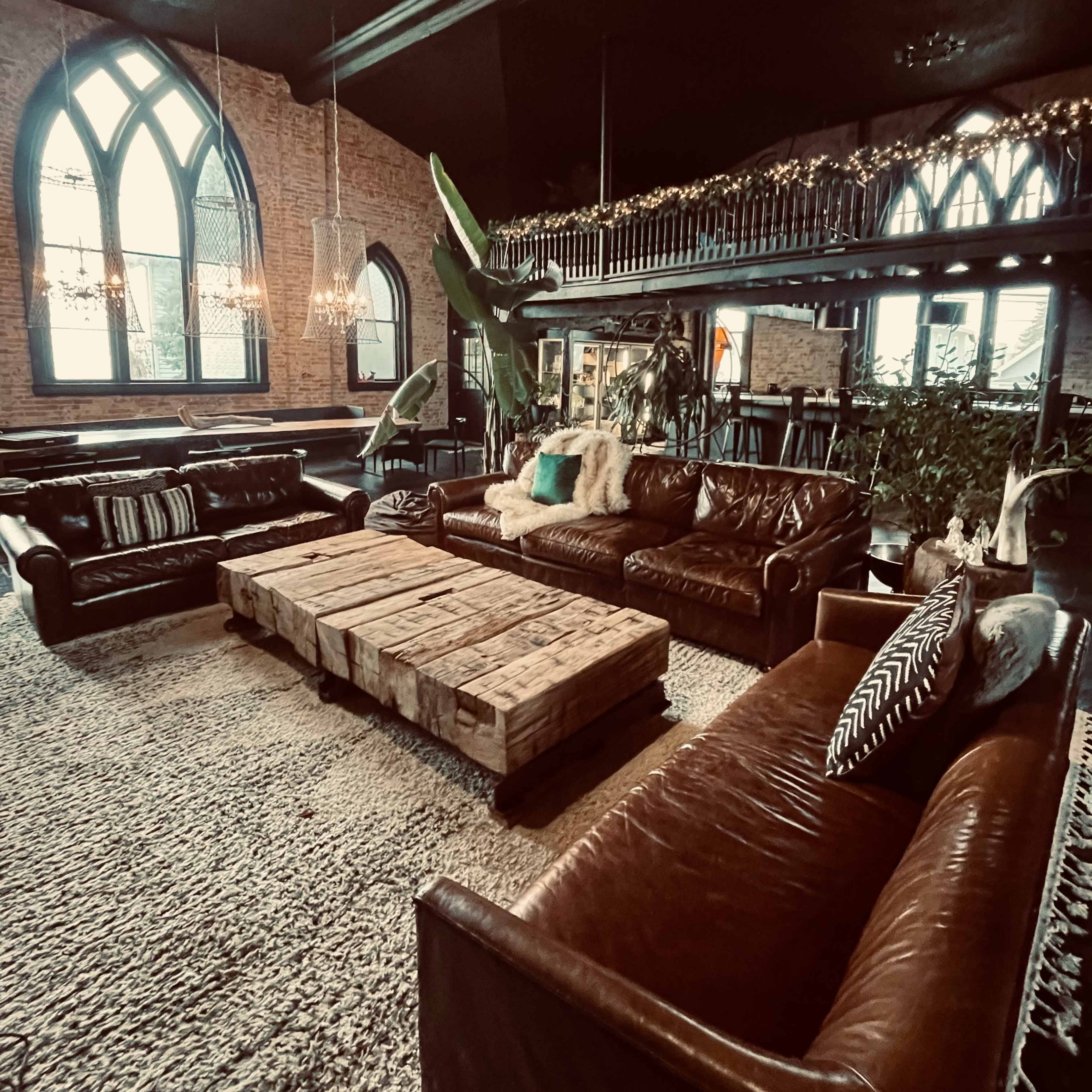 A cozy lounge area with brown leather sofas, a large wooden coffee table made from pallets, and a lush plant, set against a backdrop of brick walls and arched windows.