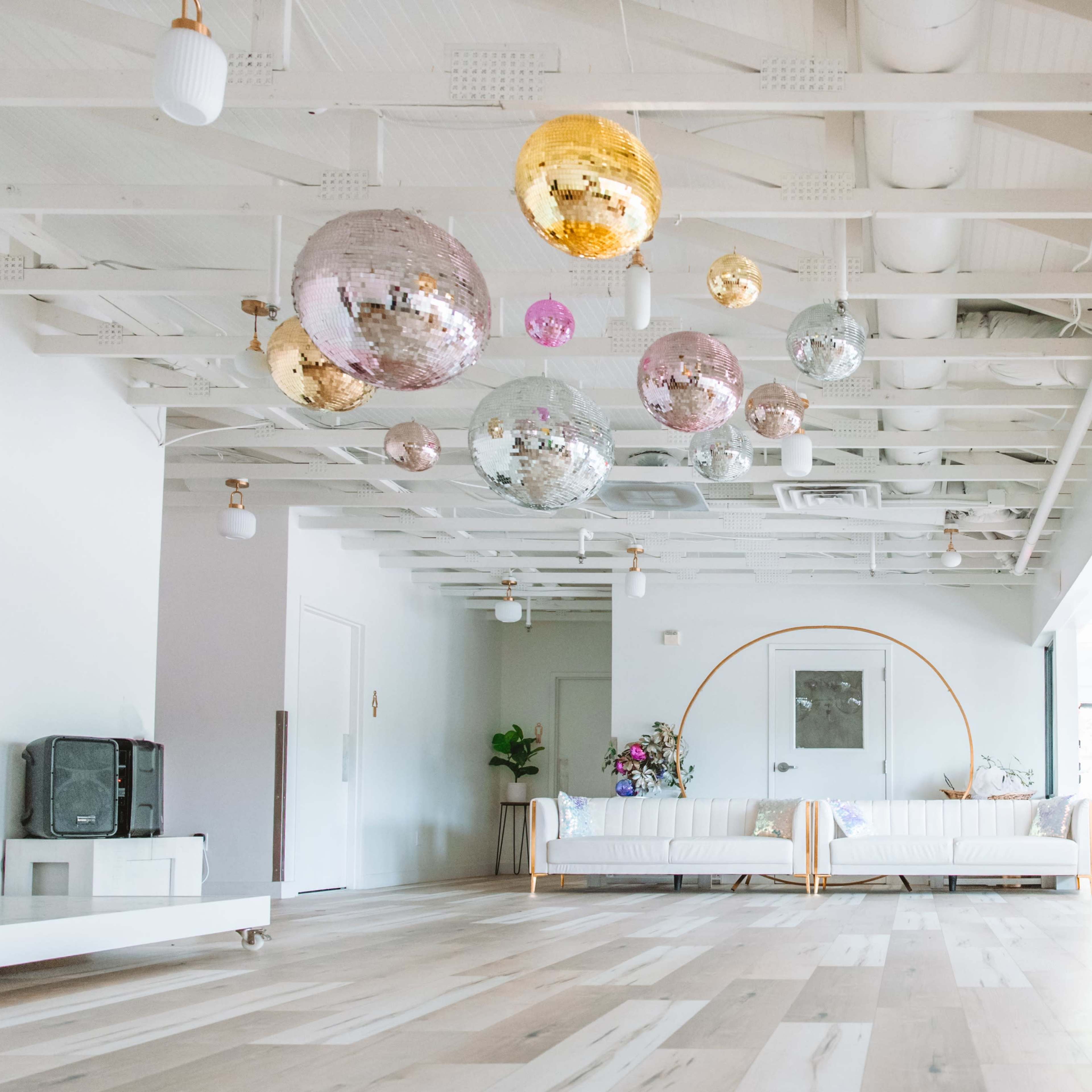 A bright, open space featuring a seating area with a sofa, decorative plants, and large hanging disco balls above hardwood flooring.
