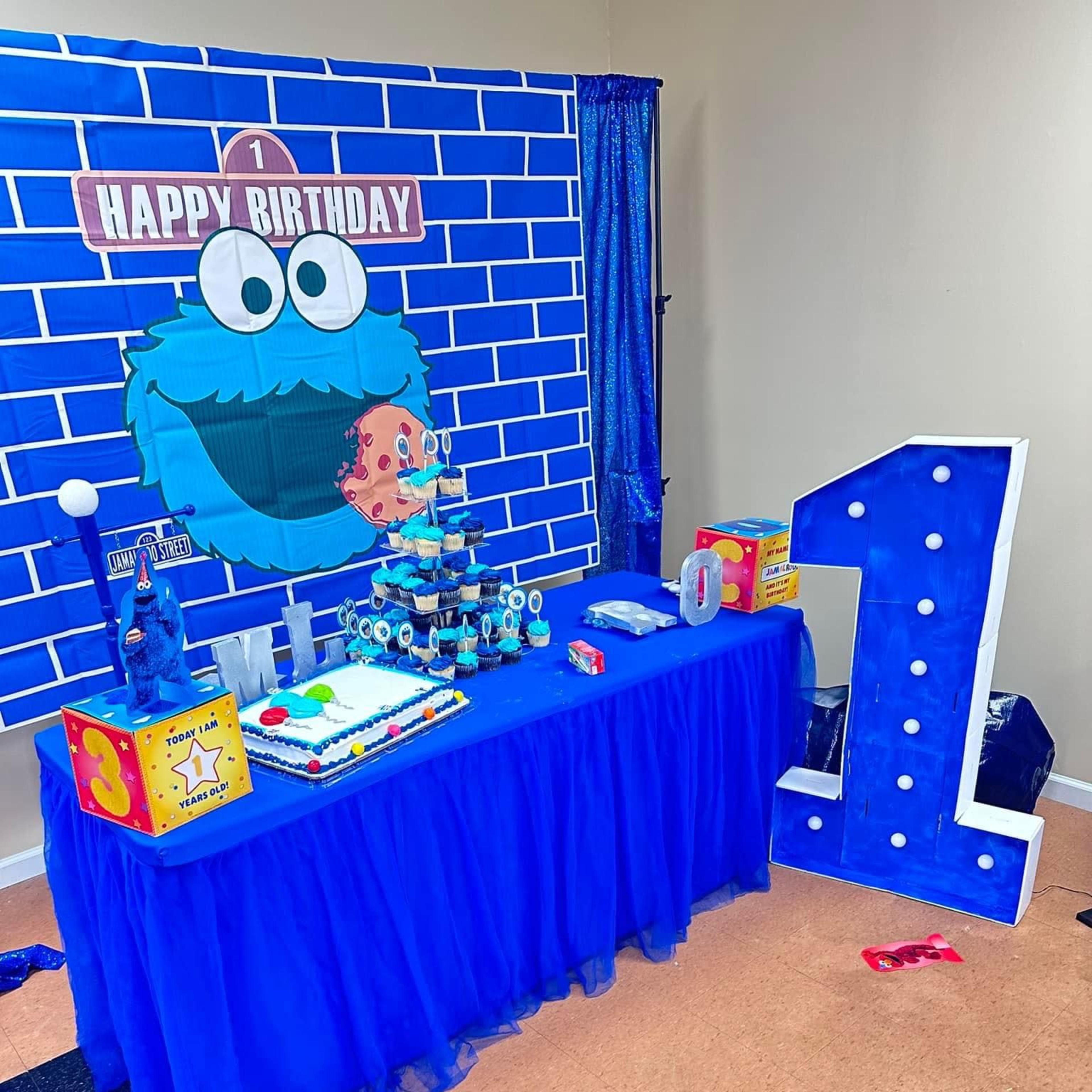 The scene shows a decorated birthday party table featuring a blue tablecloth, a cake, colorful party decorations, and a large number "1" display against a brick wall backdrop.