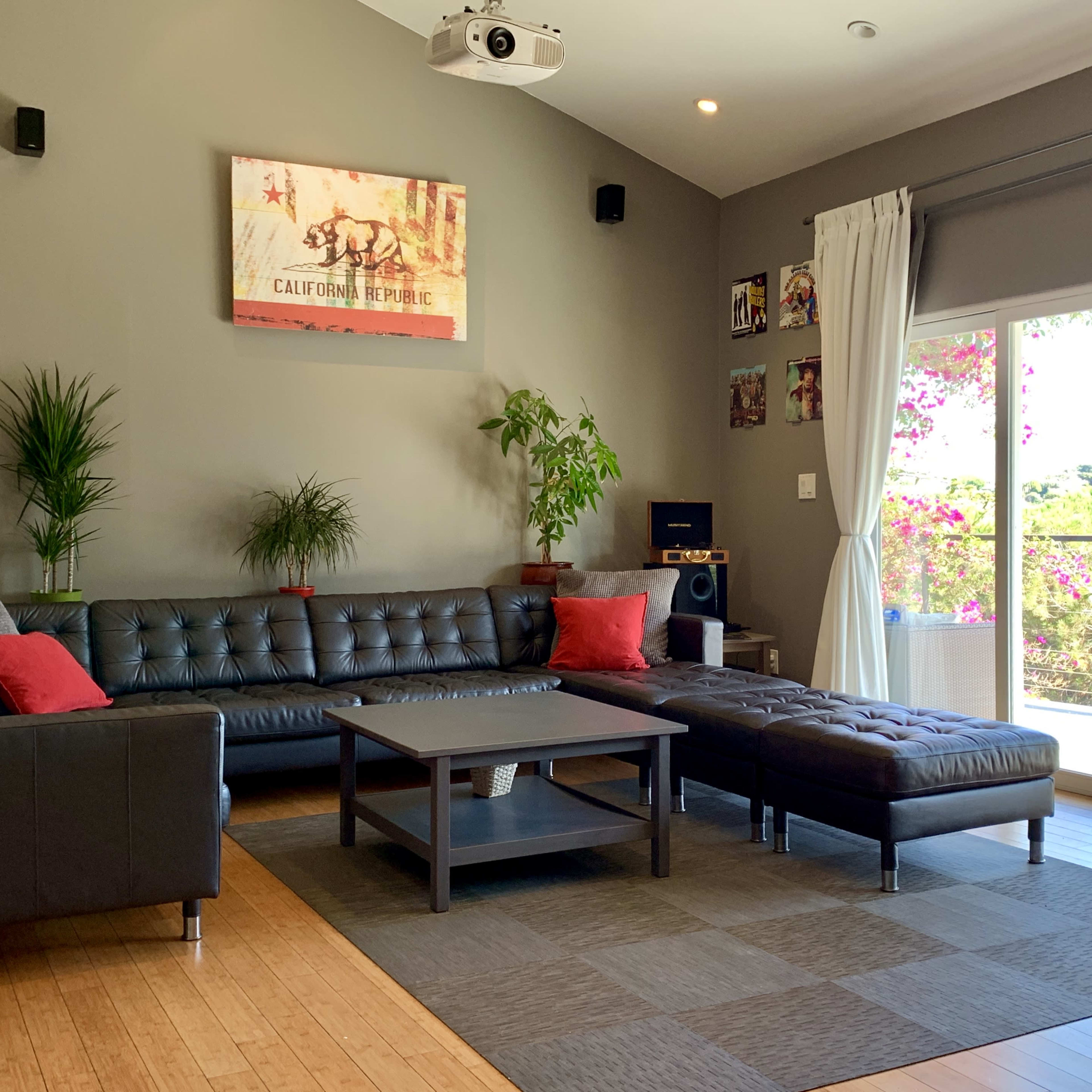 A modern living room with a large sectional sofa, a coffee table, and a projector mounted on the wall, next to sliding glass doors that open to a patio with blooming plants outside.