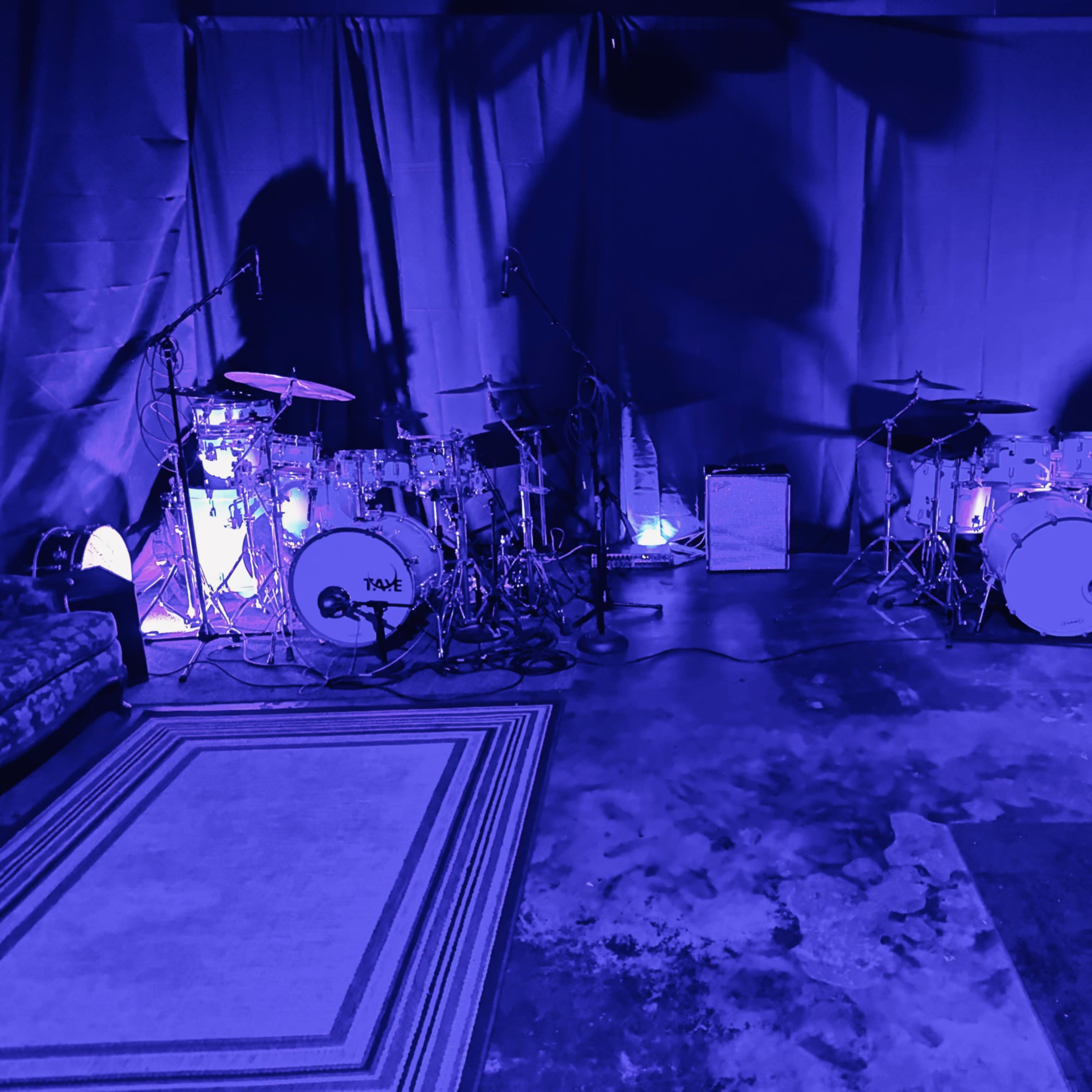 A music rehearsal space features two drum sets illuminated by purple lights, with microphones and an amplifier positioned nearby.