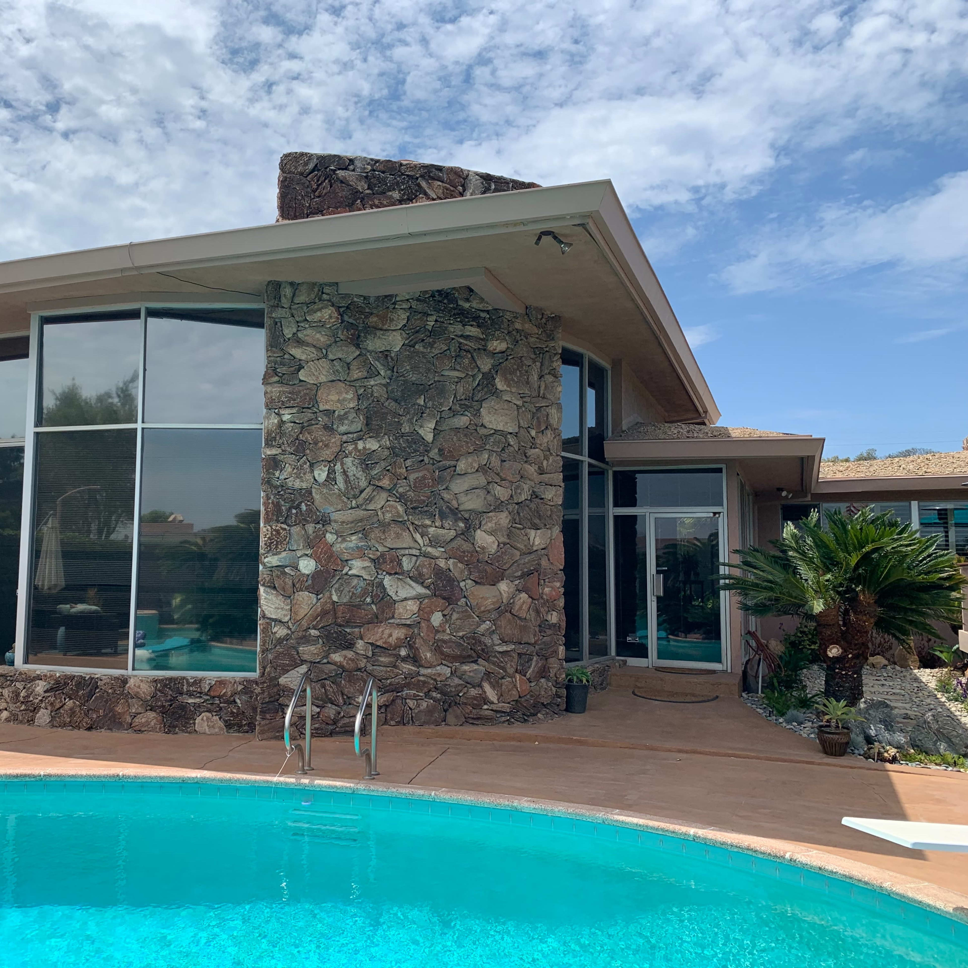 A modern house with a stone feature wall is situated next to a turquoise swimming pool under a partly cloudy sky.