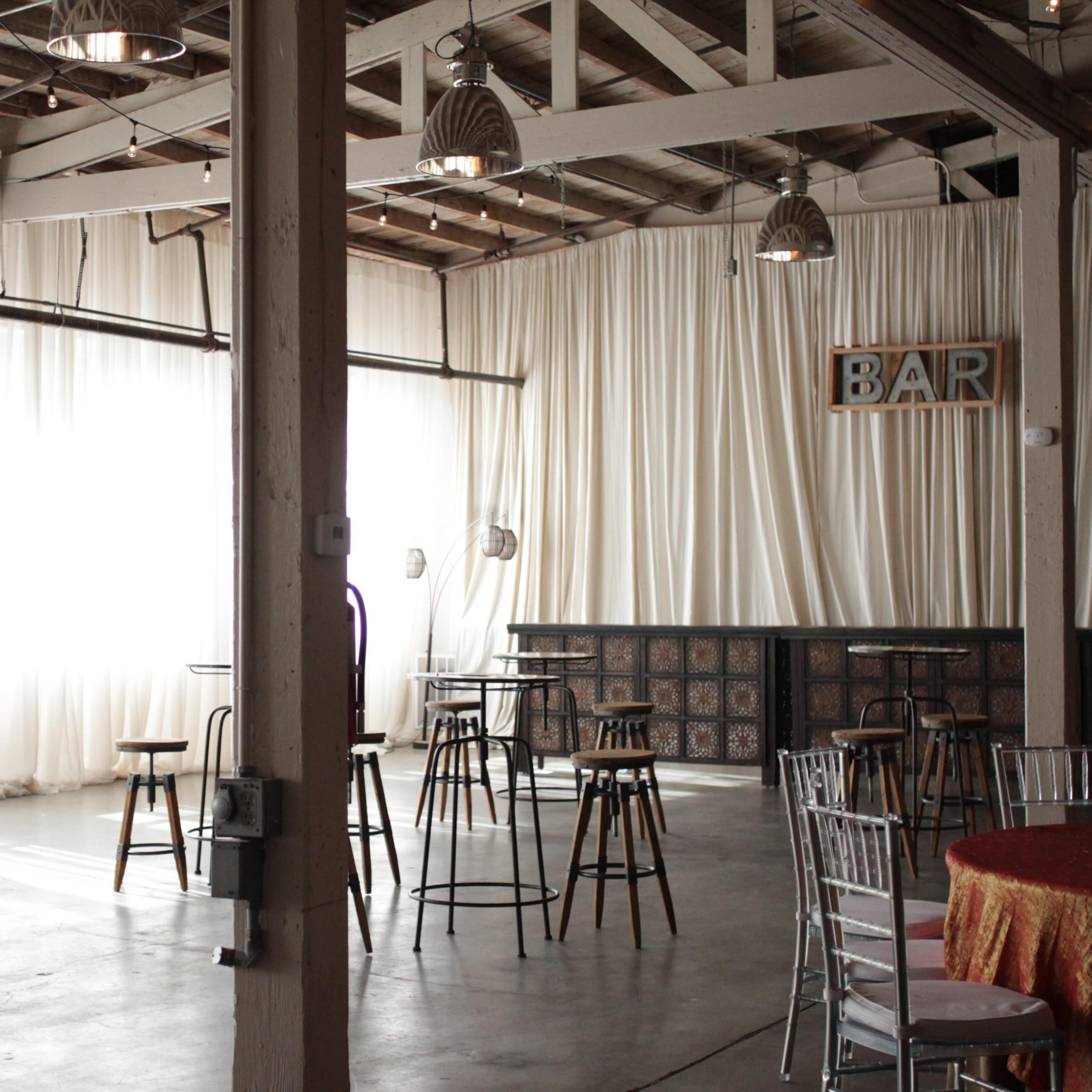 An indoor venue with tall bar tables and stools arranged among large windows draped with sheer curtains, alongside a bar area.