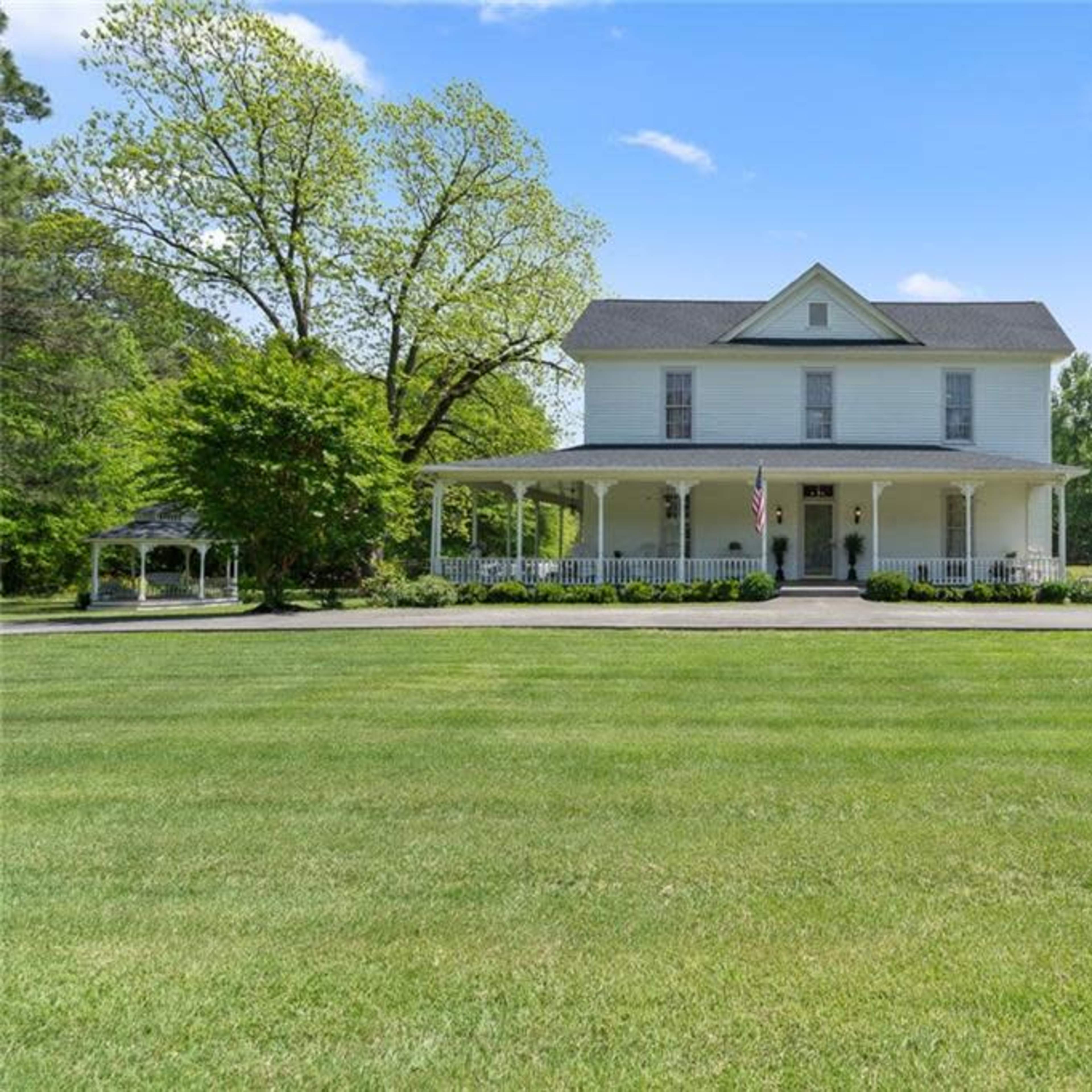 A large, two-story yellow house with a porch is set in a spacious, green yard surrounded by trees.