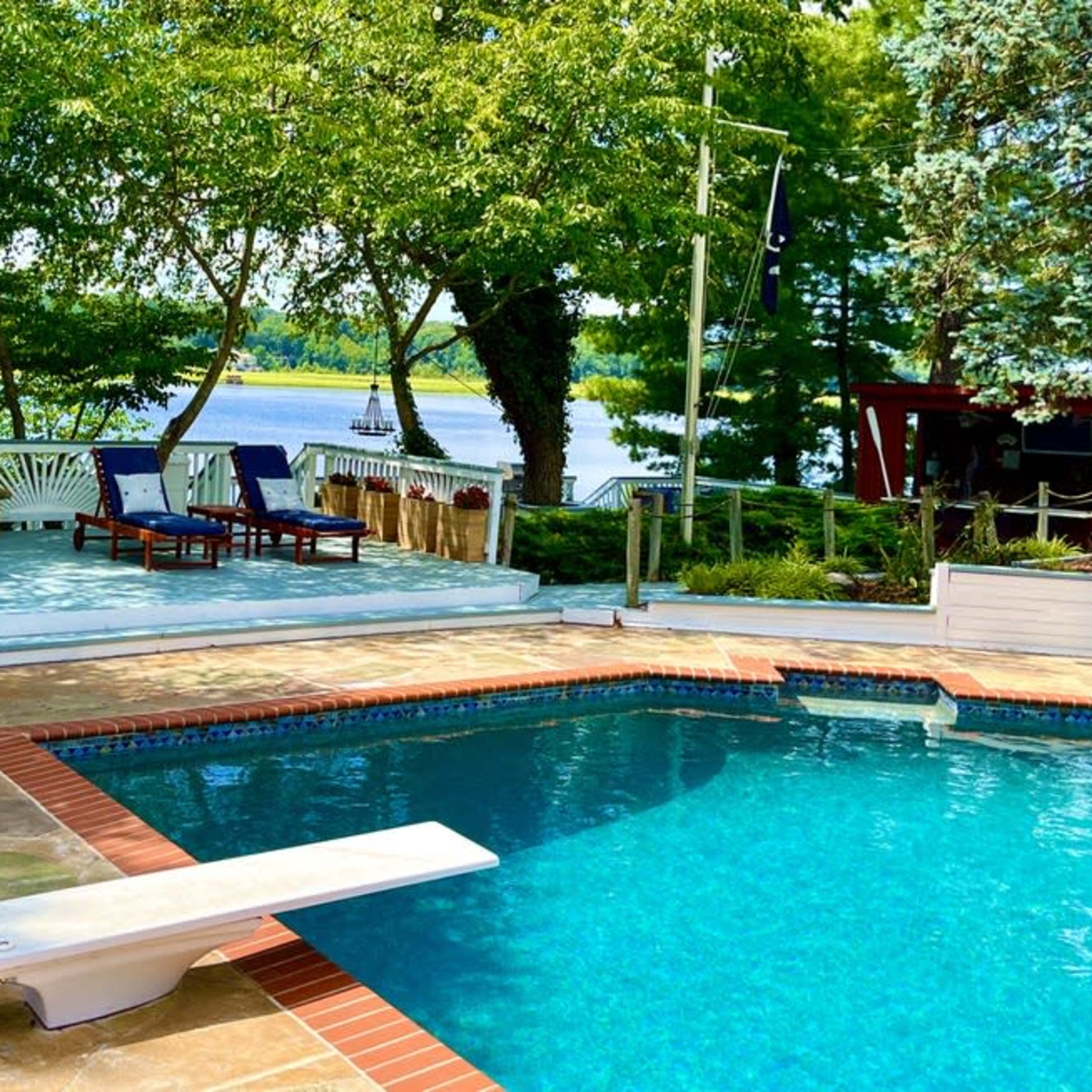 A backyard area featuring a swimming pool, a diving board, and a patio with seating that overlooks a lake surrounded by trees.