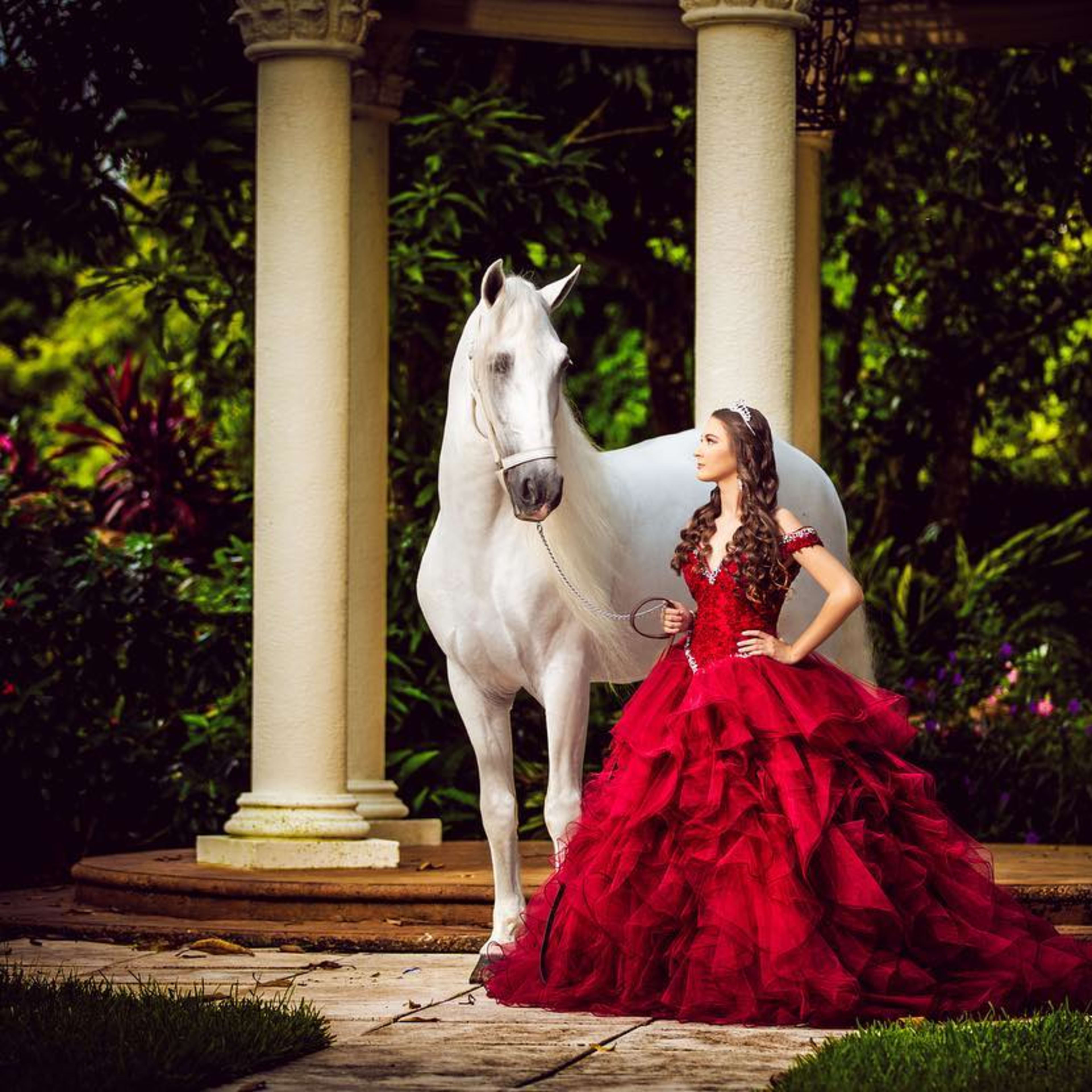 A young woman in a flowing red gown stands beside a white horse near a columned structure in a lush garden.