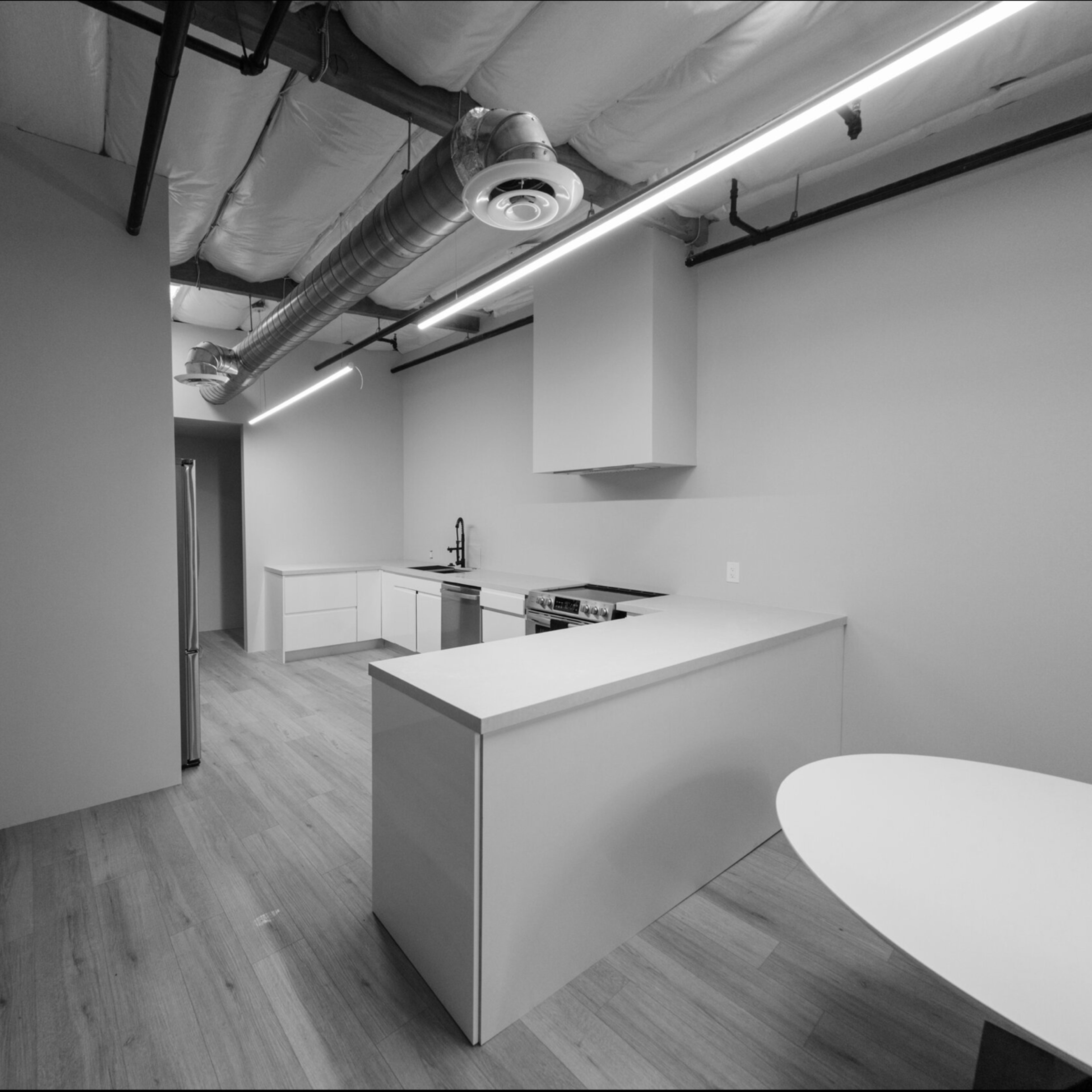 A spacious, minimalistic kitchen area featuring a central island, white cabinetry, and modern appliances, with a light wood floor and exposed ceiling ducts.