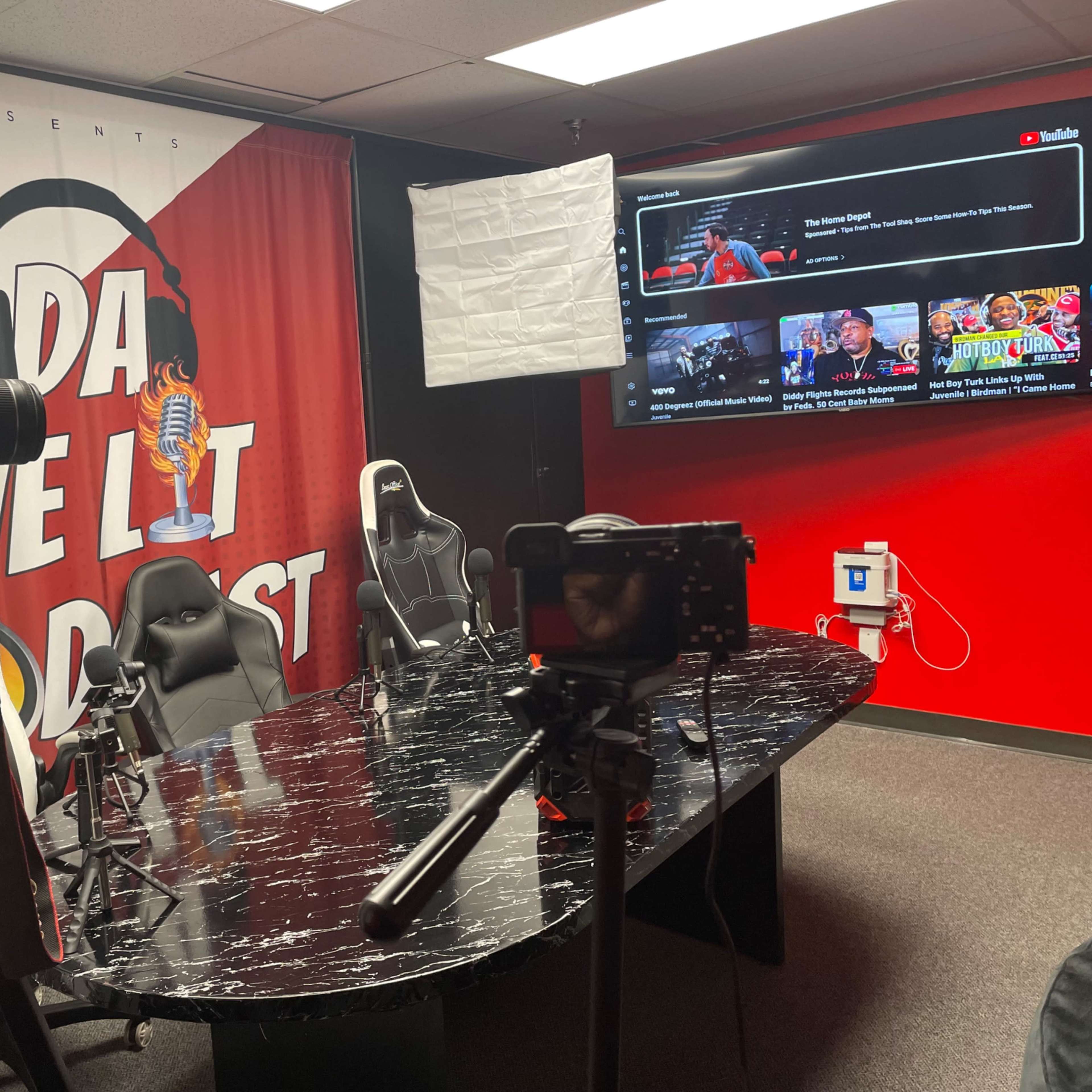 A recording studio with a large, round table surrounded by gaming chairs, cameras on tripods, and a backdrop displaying the title "DA WE LIT PODCAST."