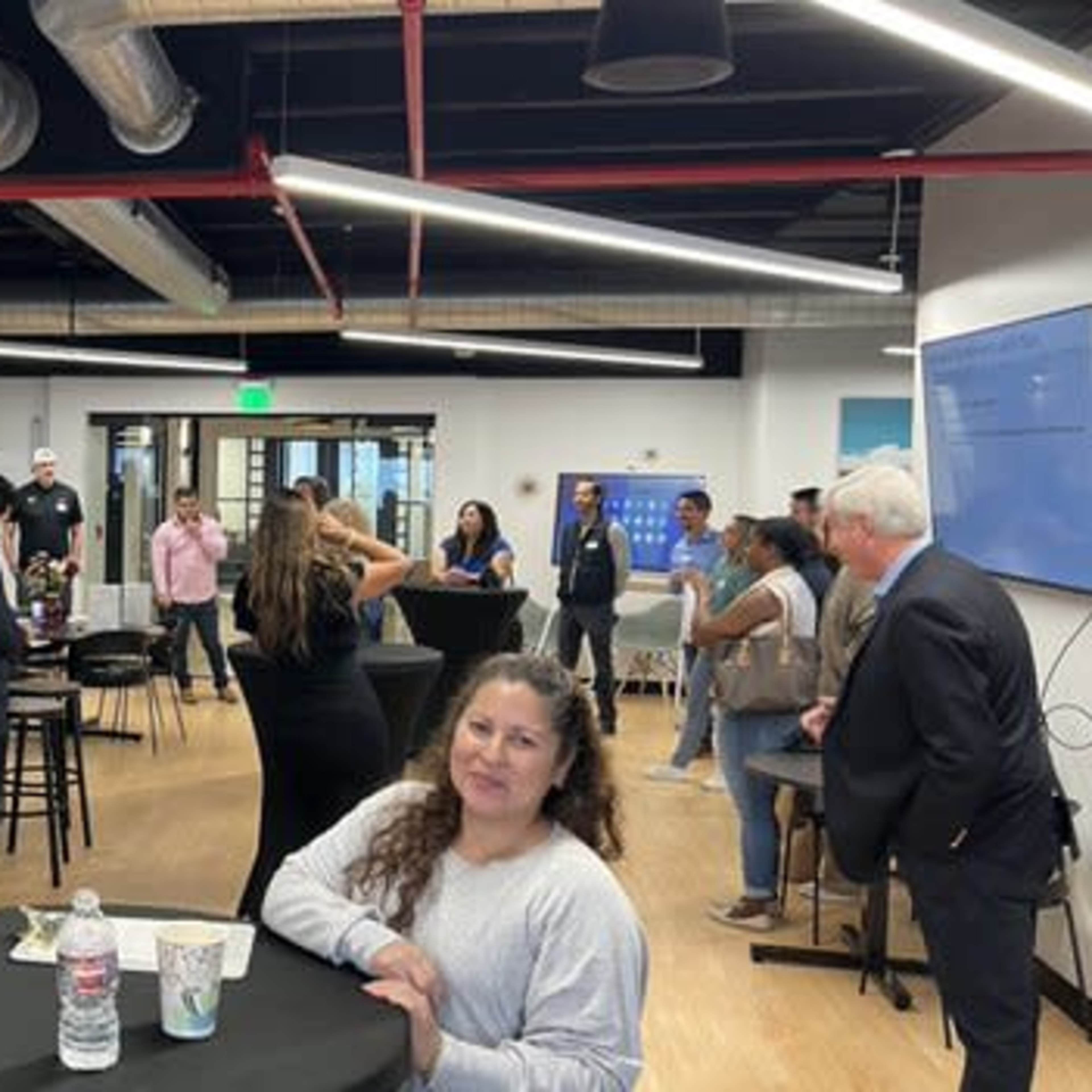 A group of people are gathered in a modern office space with high tables and a bar area, engaging in conversation during an event.