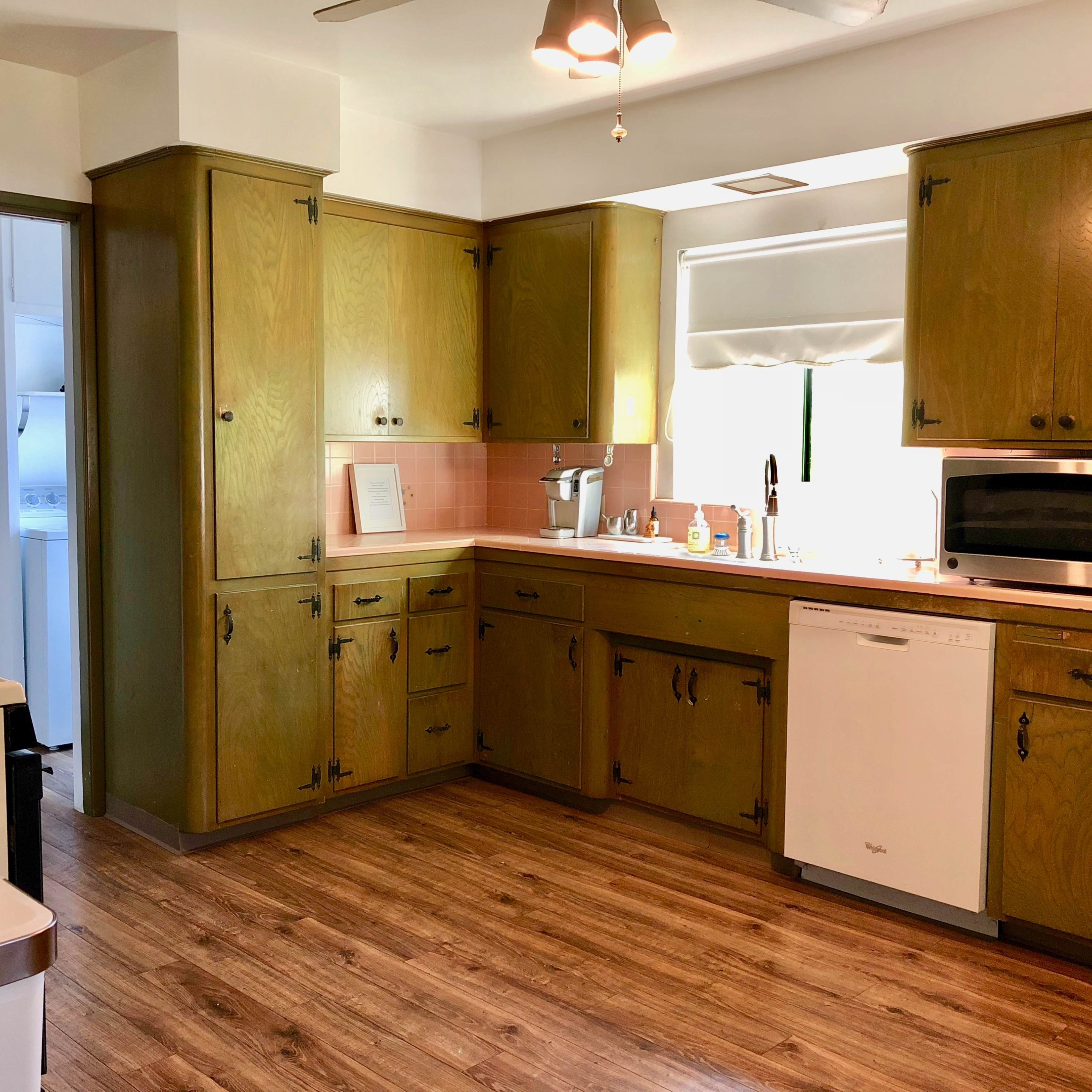 A kitchen with wooden cabinet doors, a sink, a microwave, and a dishwasher, featuring light pink tiled walls and wood flooring.