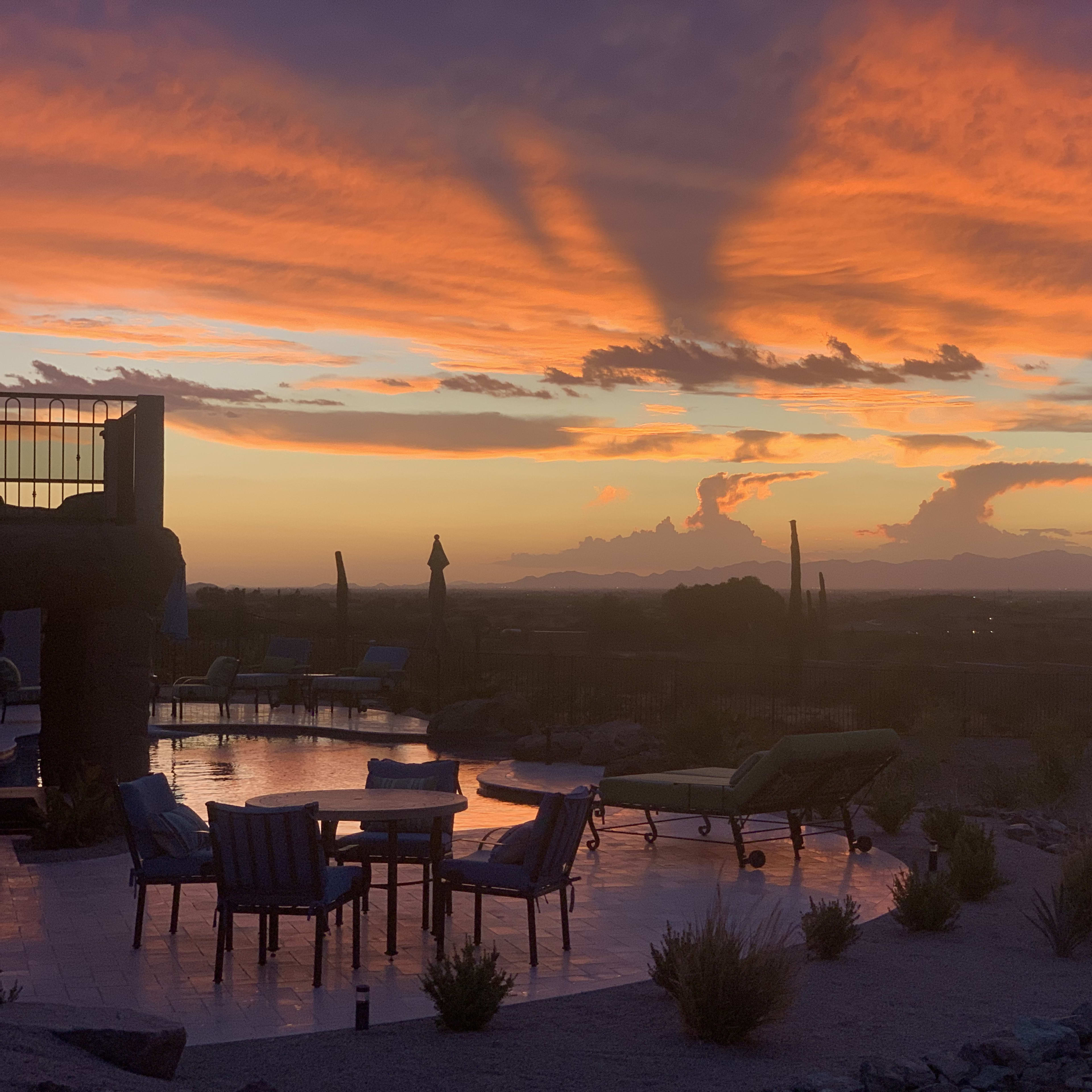 A sunset casts vibrant colors across the sky, reflecting off a pool surrounded by seating and desert vegetation.
