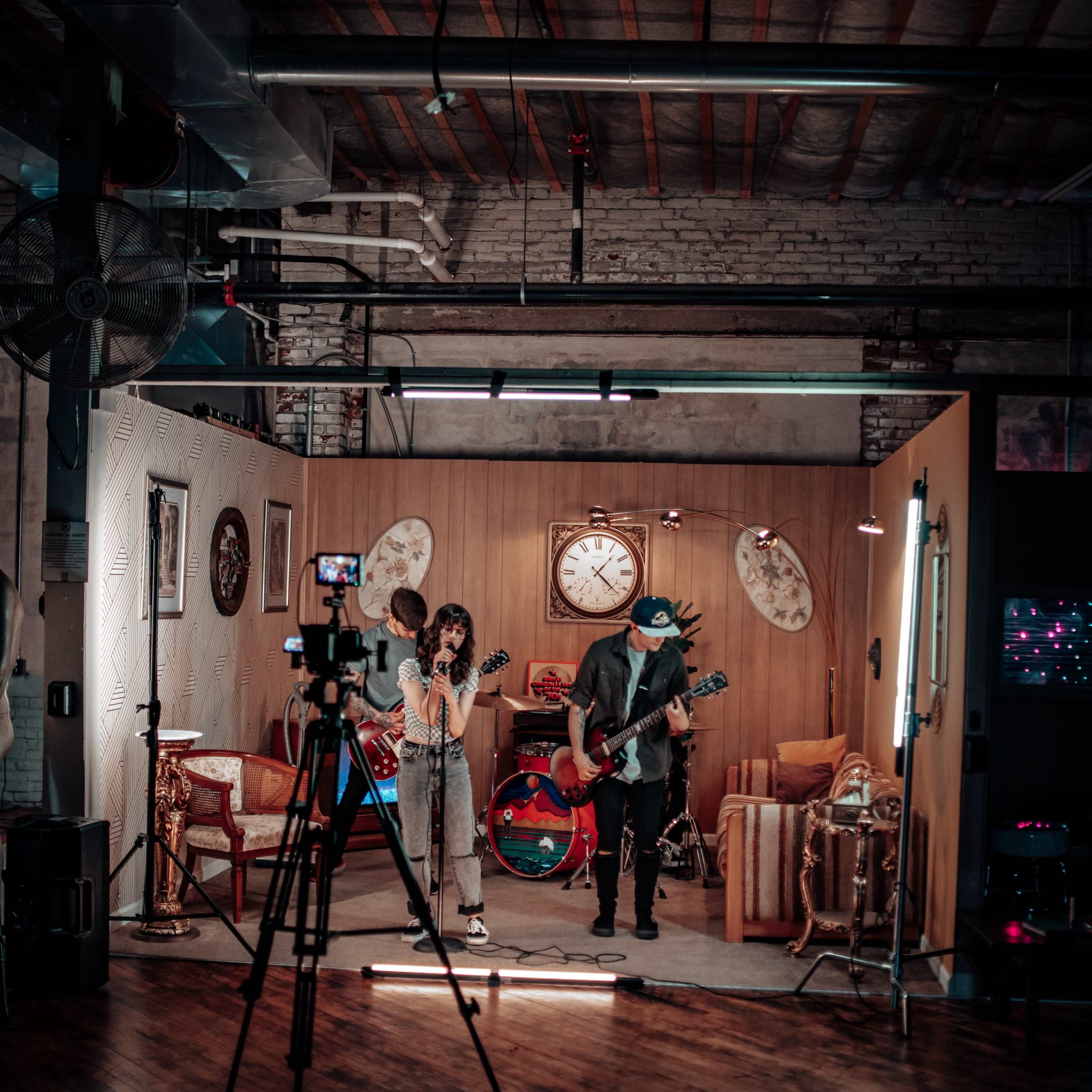 A band performs in a studio setting with wooden floors and retro decor, including clocks and furniture, while a camera is positioned to record the performance.