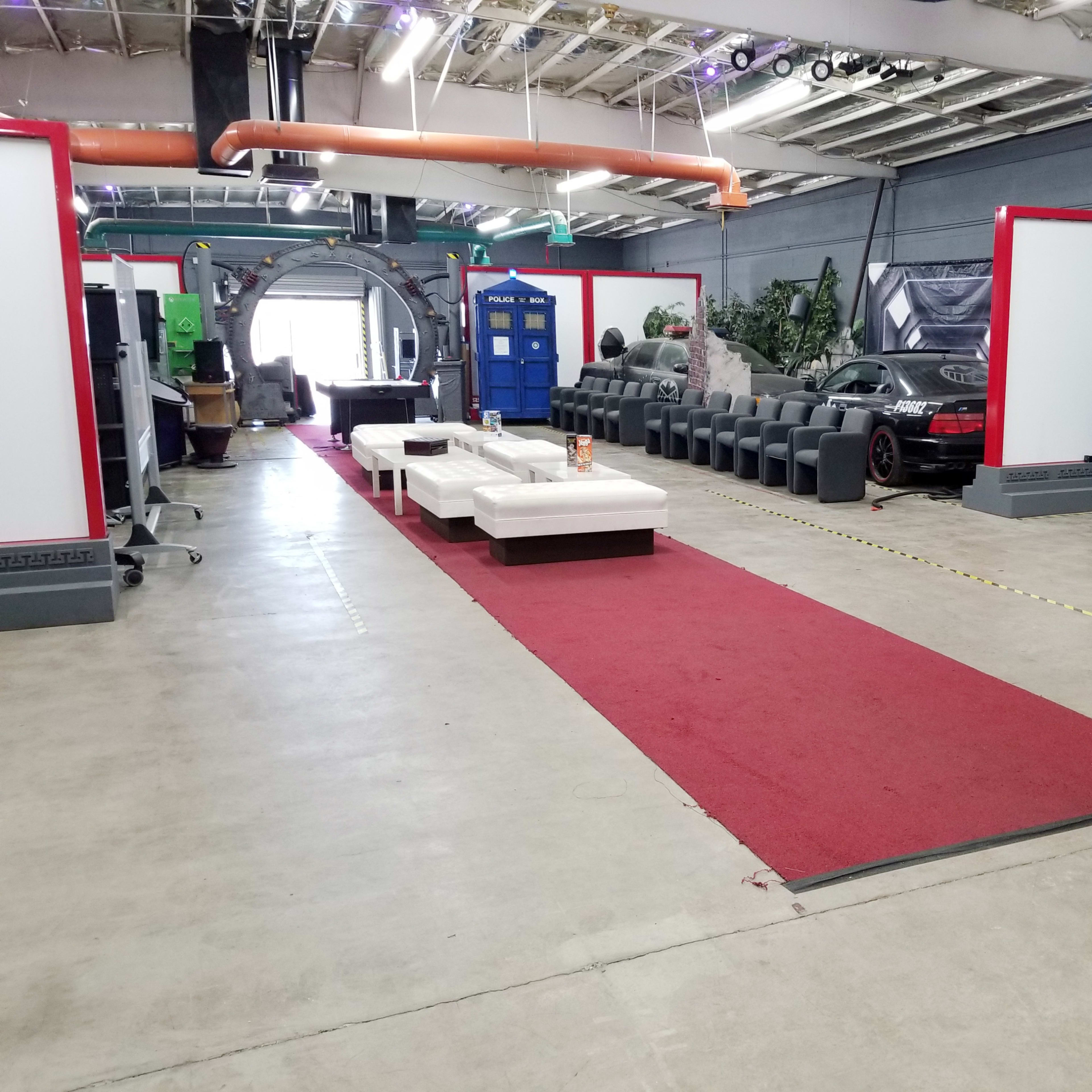 A spacious indoor area with a red carpet leading through rows of seating and a TARDIS, alongside a parked car and recycling bins.