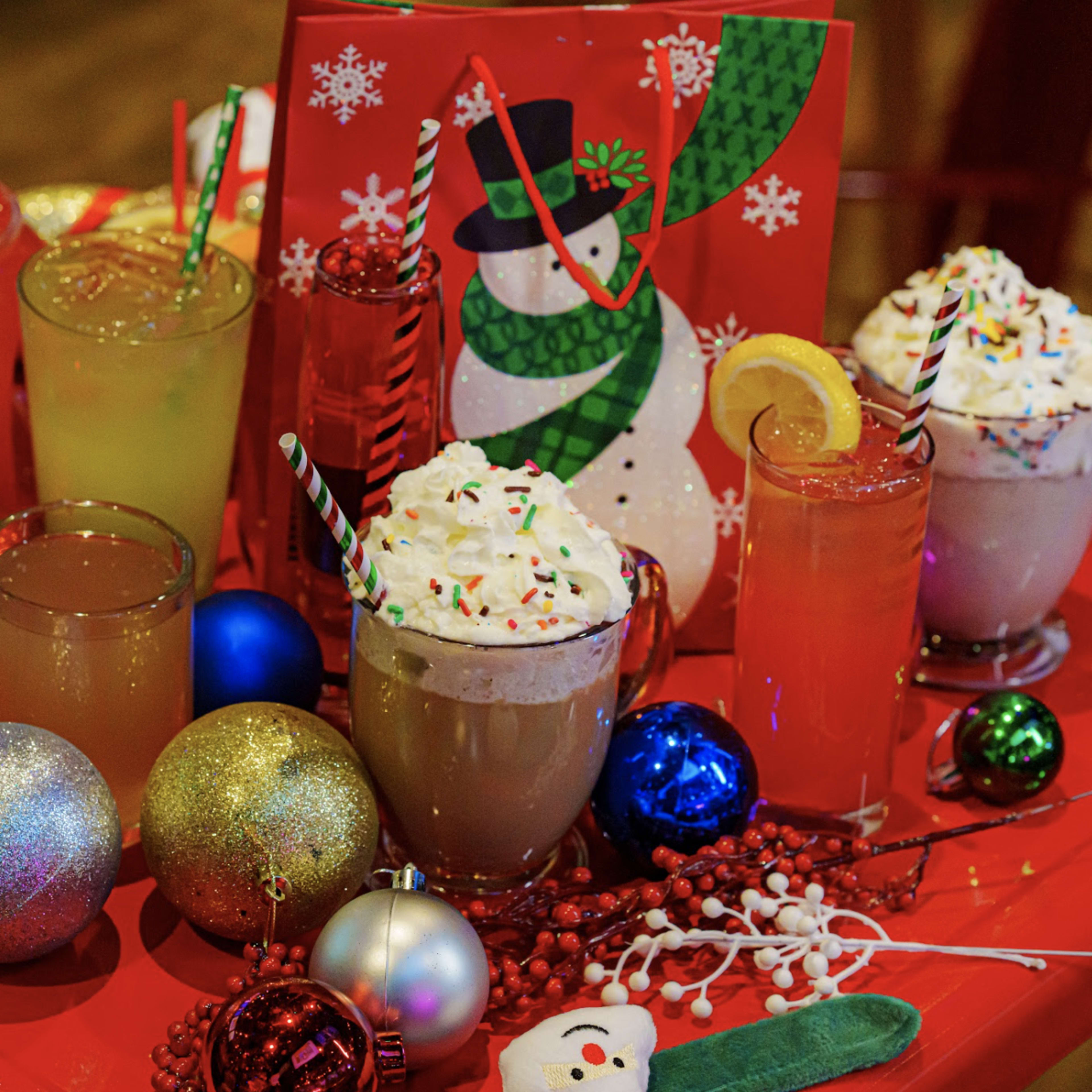 A festive display of various colorful holiday drinks surrounded by Christmas decorations, including ornaments and a snowman-themed card.