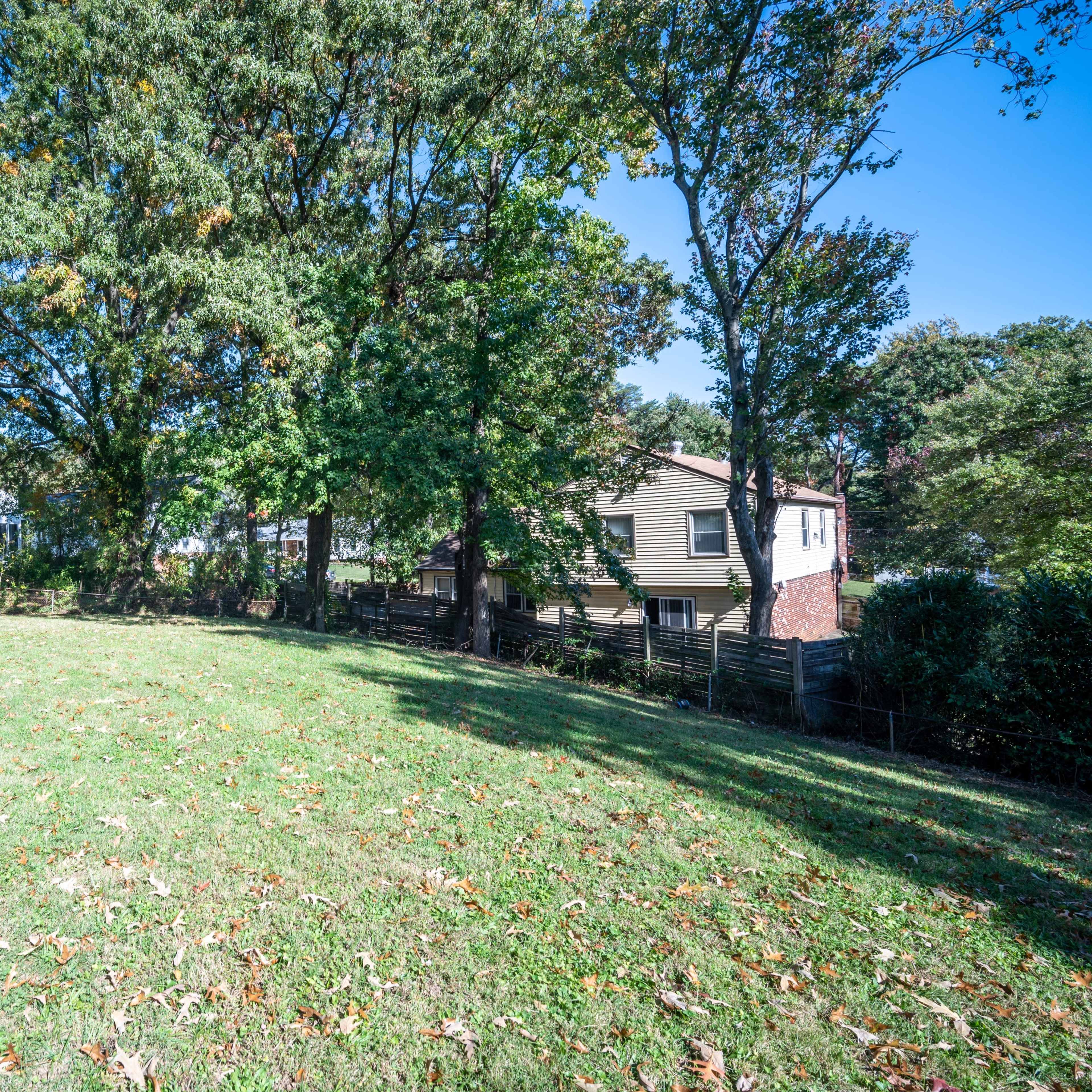 A grassy backyard is bordered by trees and nearby houses under a clear blue sky.
