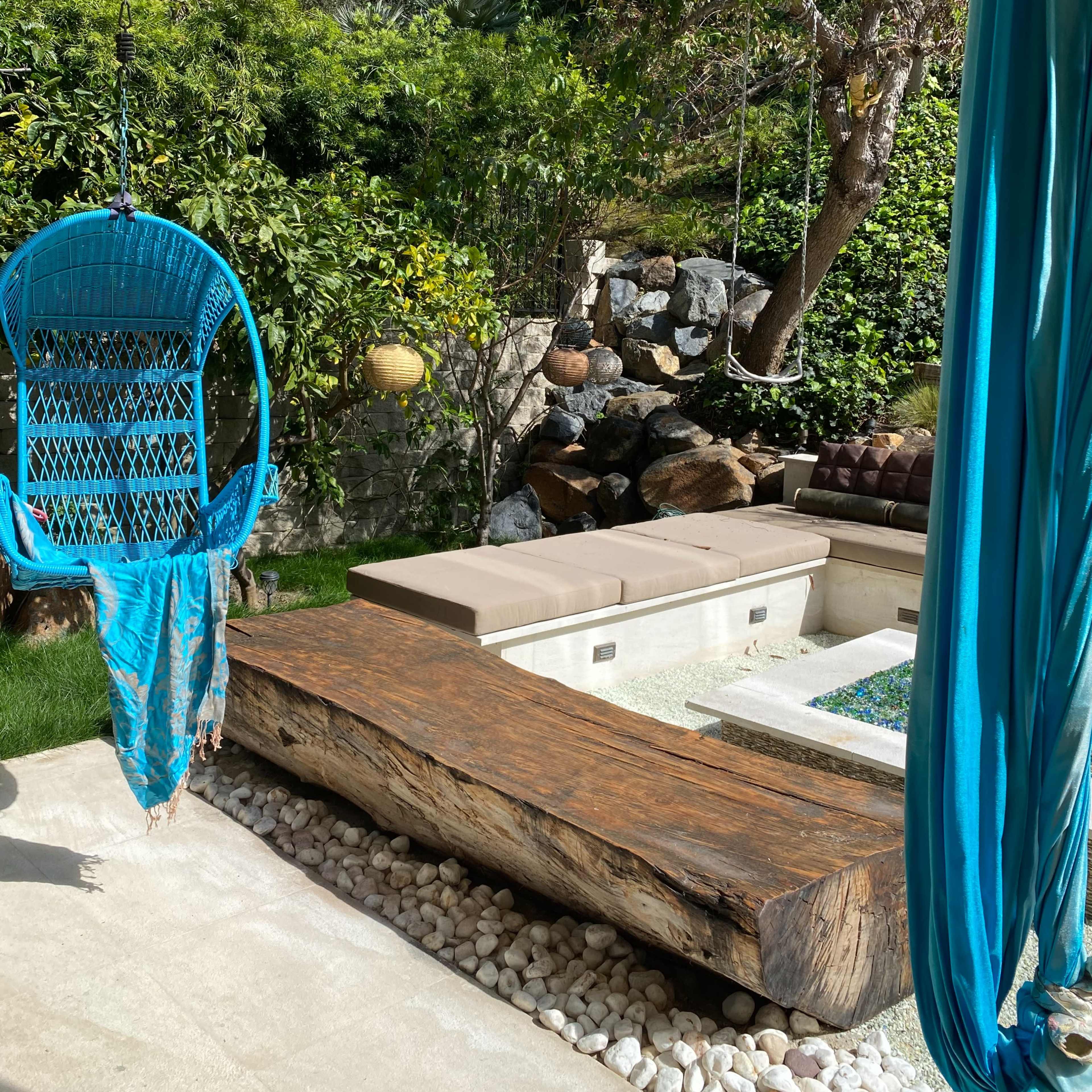 A turquoise swing chair hangs beside a wooden bench and a central water feature in a lush garden.