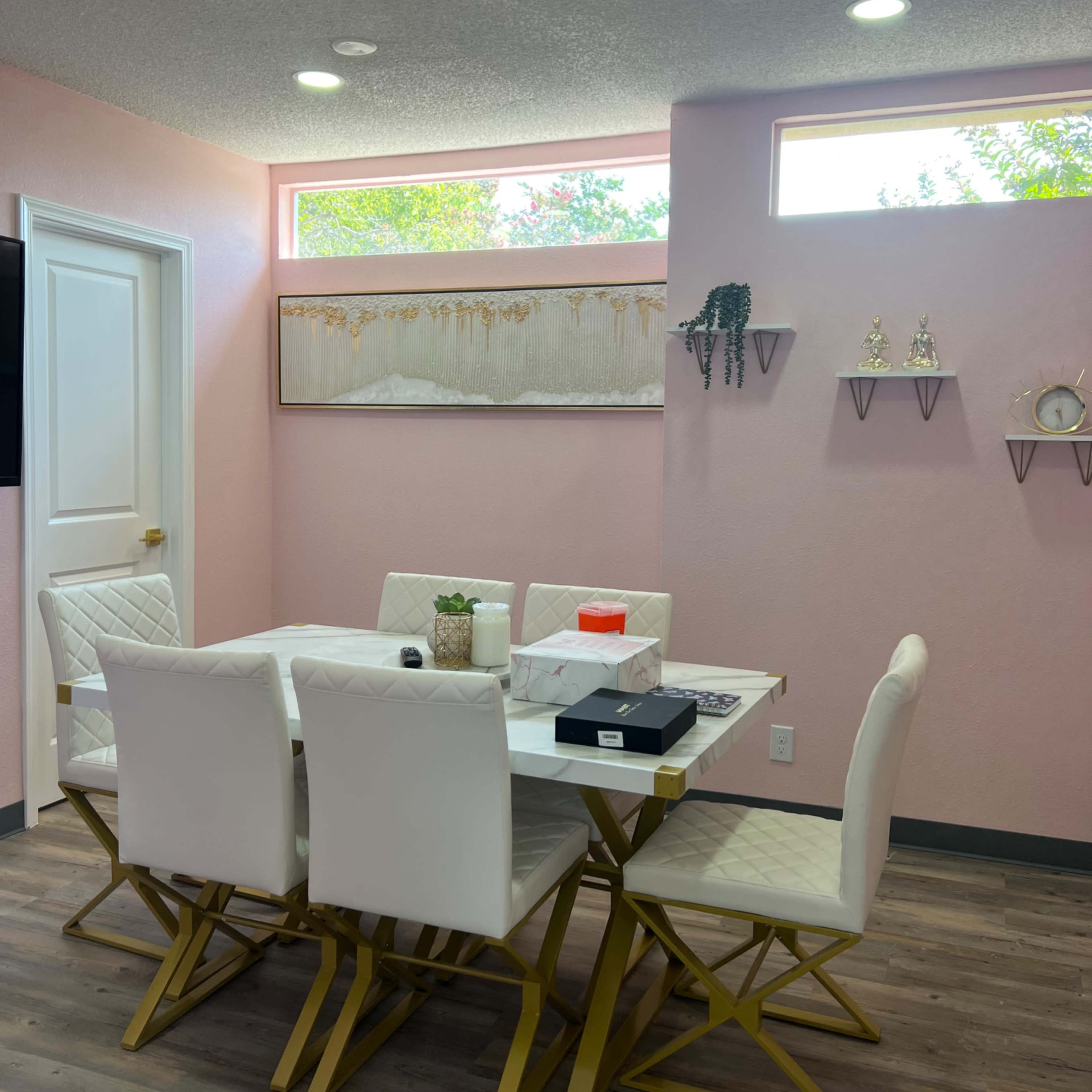A dining table with white chairs and a gold base is positioned in a brightly lit room with pink walls and windows, featuring decorative wall shelves and artwork.