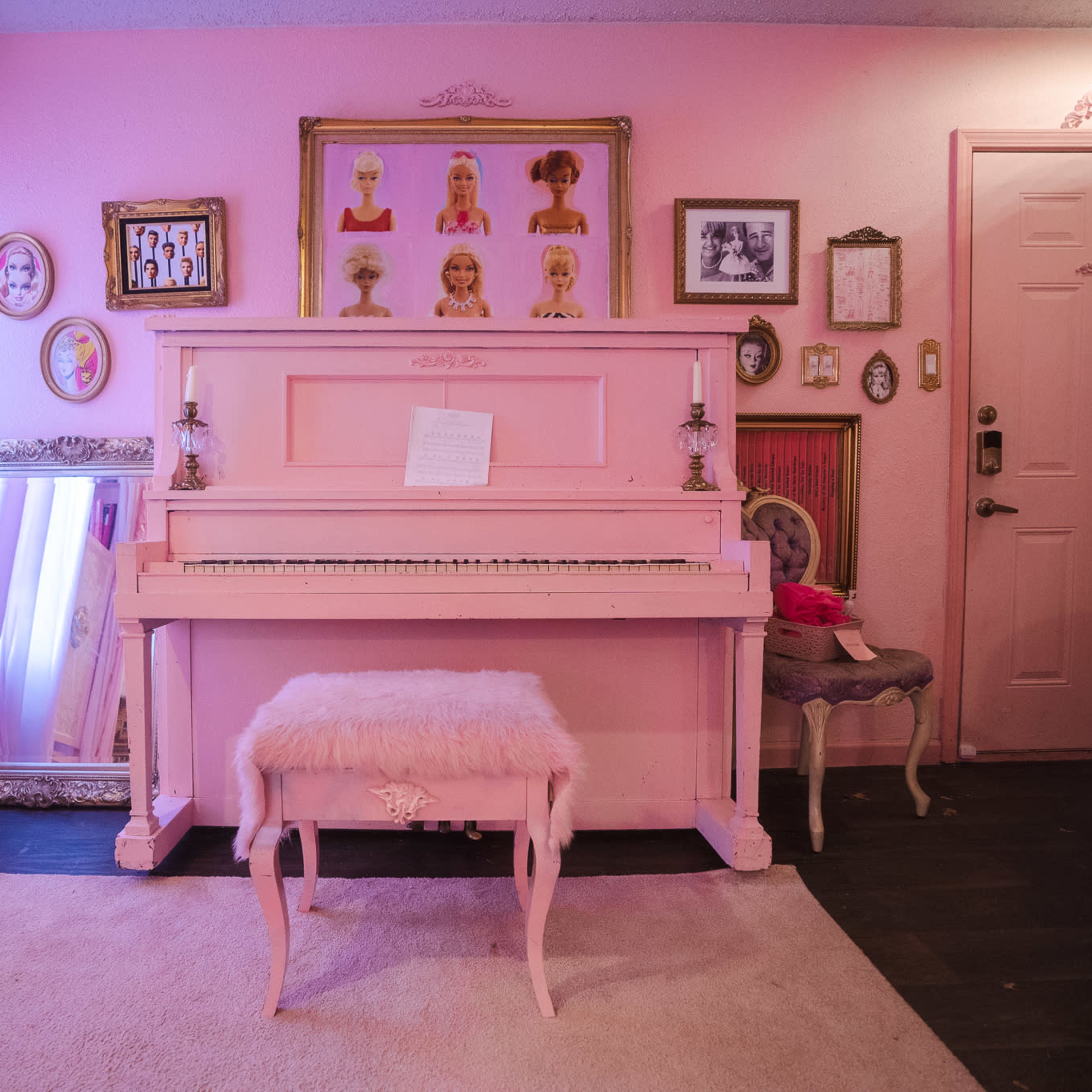 A pink piano is positioned against a wall adorned with framed portraits and decorative mirrors in a room with light pink curtains and a plush rug.
