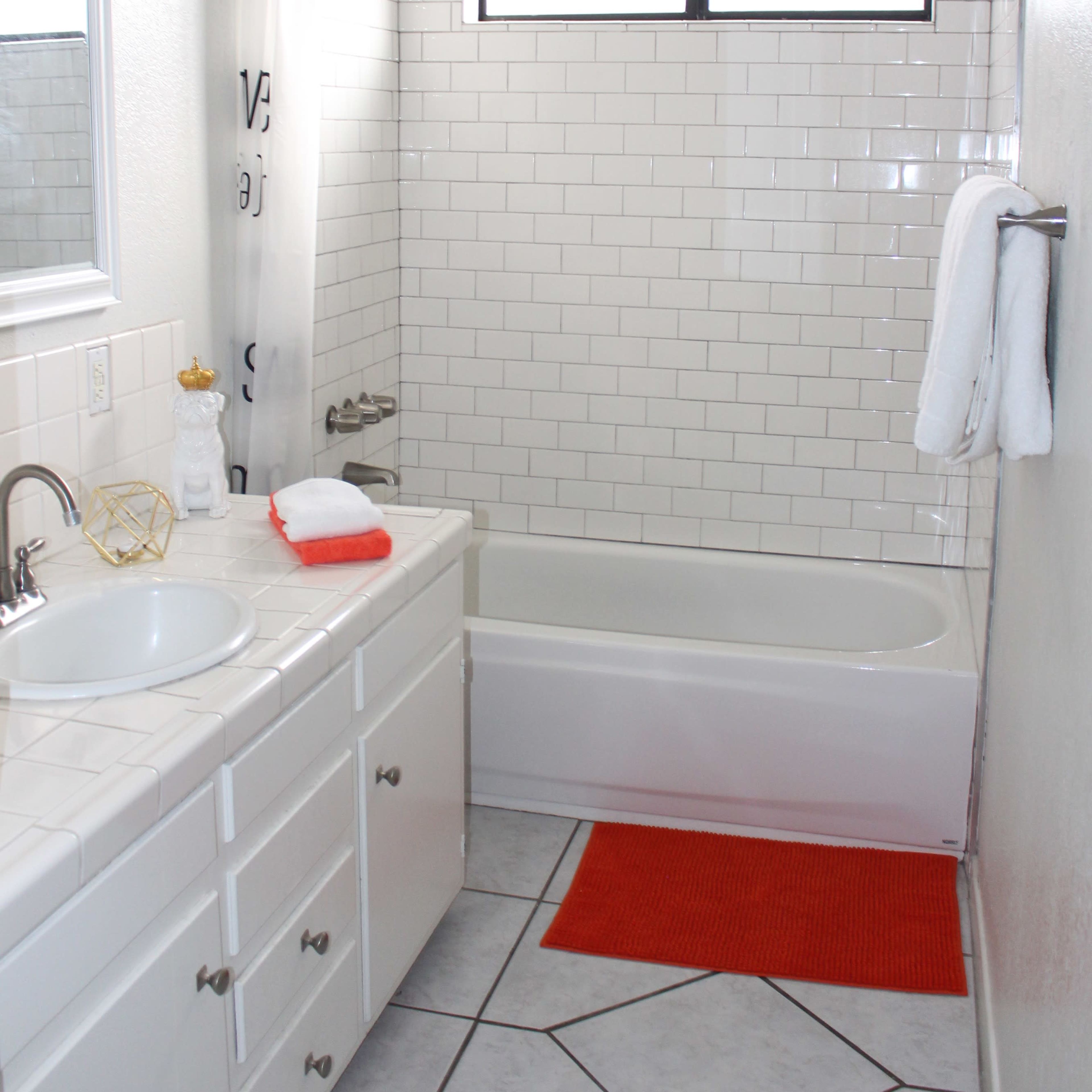 A clean bathroom featuring a bathtub with a showerhead, a white sink with a mirror, and a bright orange bath mat on the tiled floor.