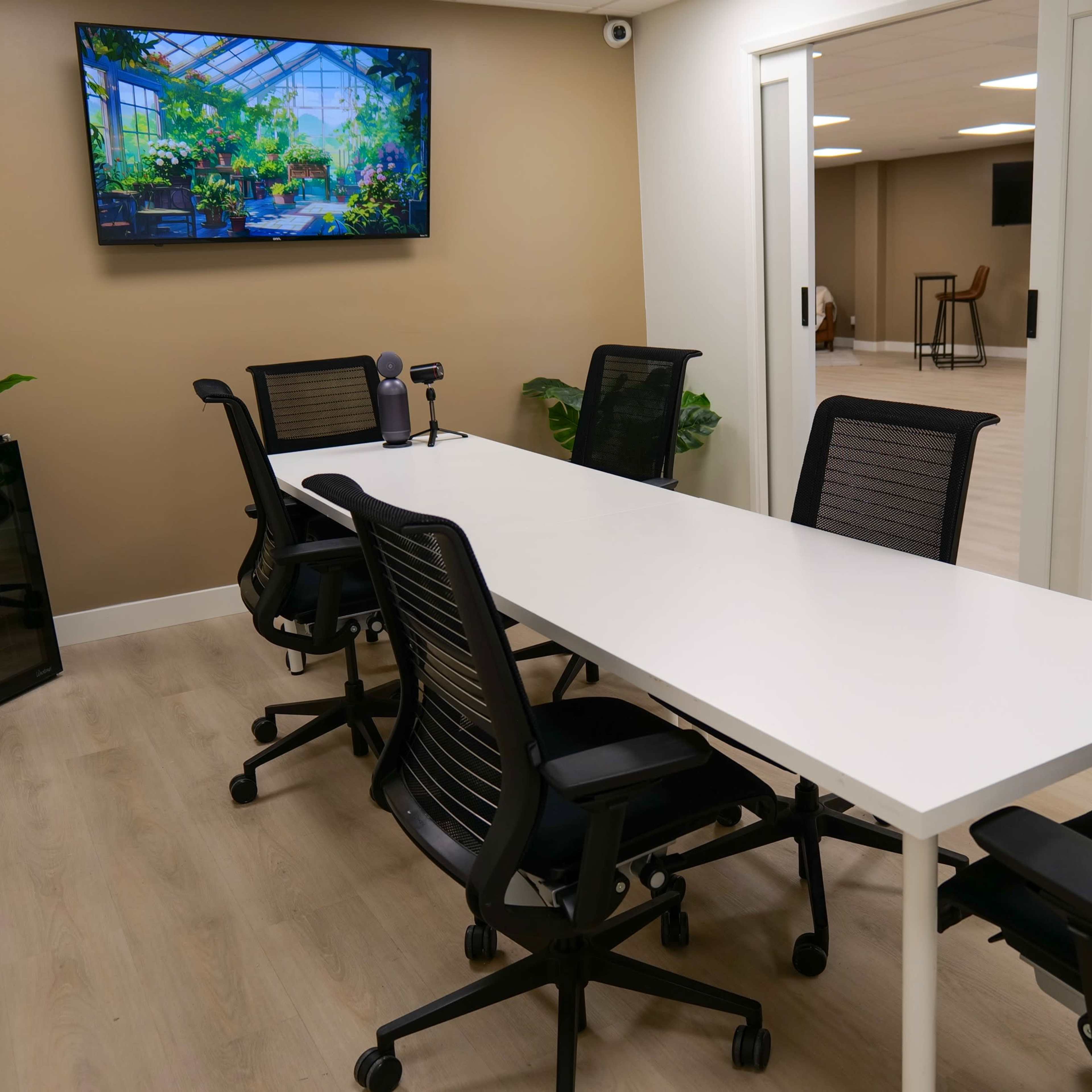 A modern conference room features a long white table surrounded by black ergonomic chairs, with a large screen displaying greenery on the wall.