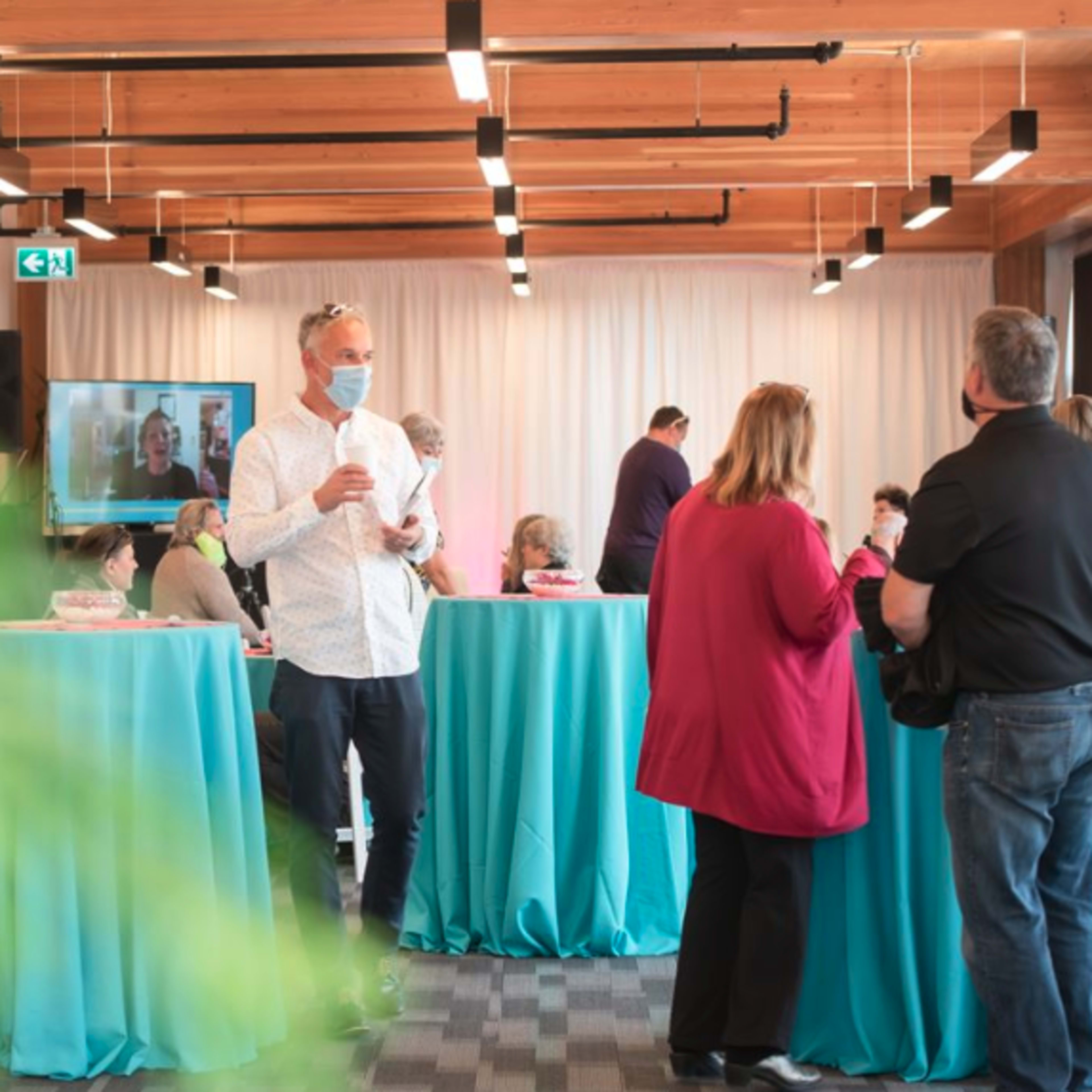 A group of people stands around teal tables in a room with wooden beams, some engaging in conversation while others watch a video presentation on a screen.