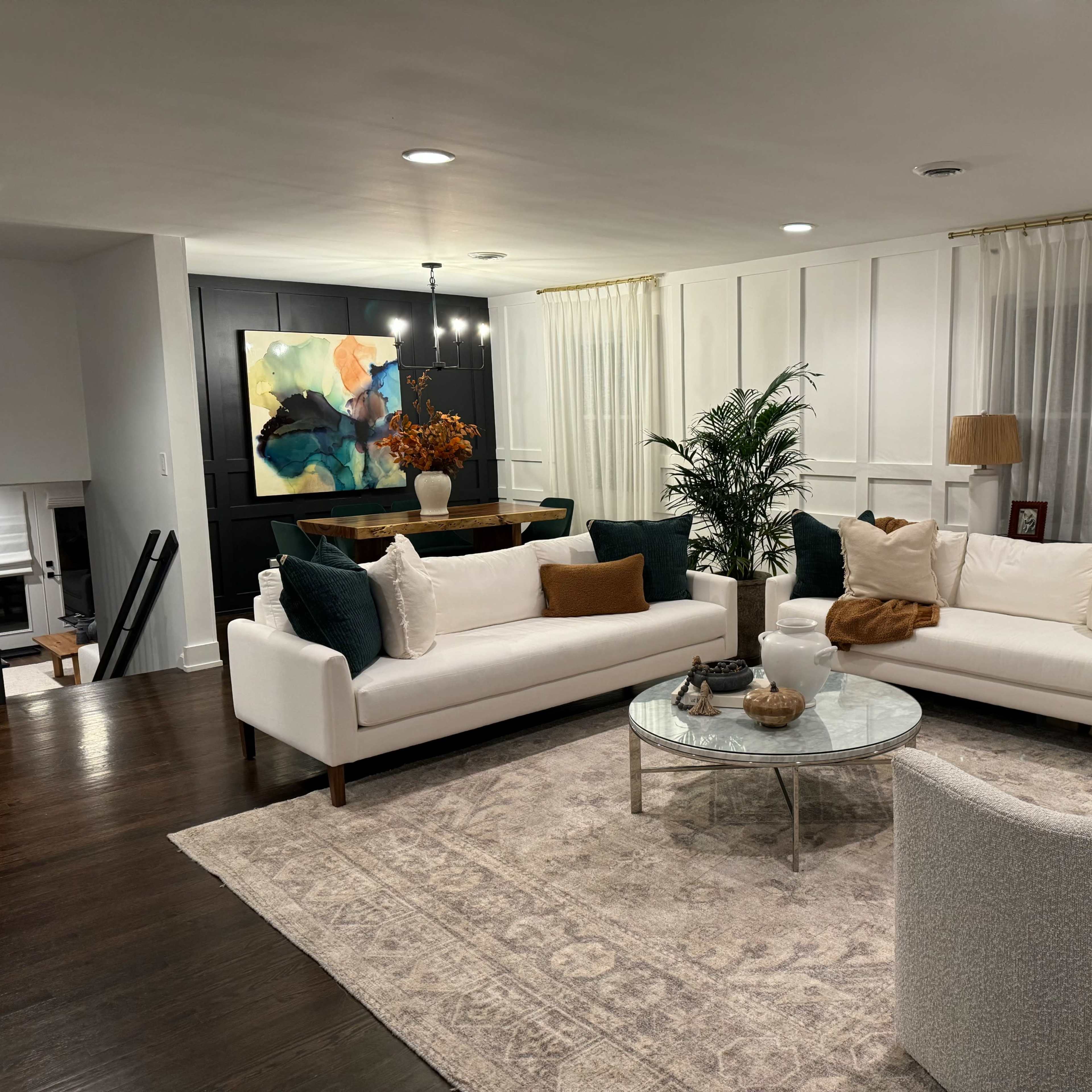 A modern living room featuring a white sectional sofa and armchairs, a round glass coffee table, and a large abstract painting on the wall.