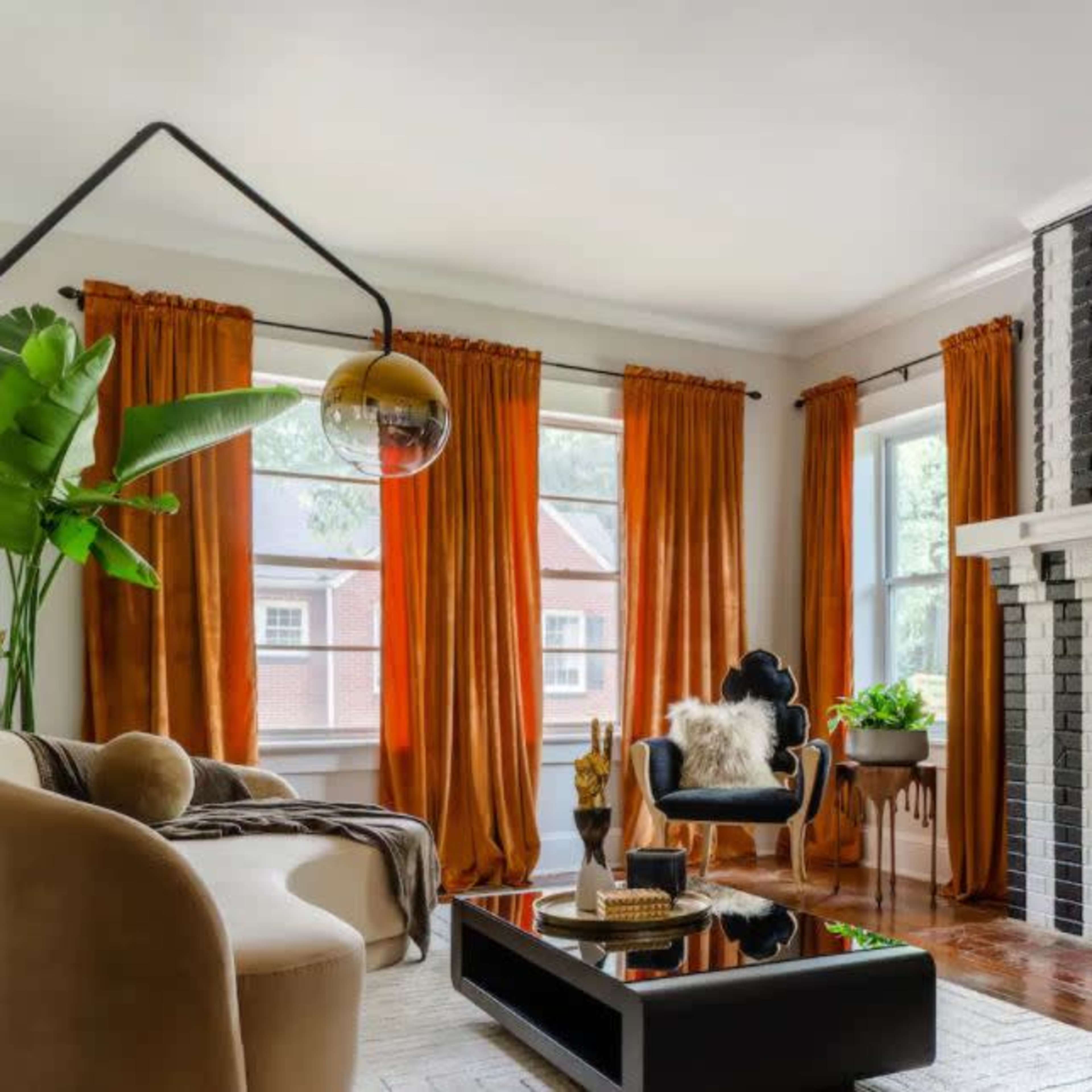 The living room features a black and white striped fireplace adorned with stars, complemented by large orange curtains, a beige sofa, and potted plants.