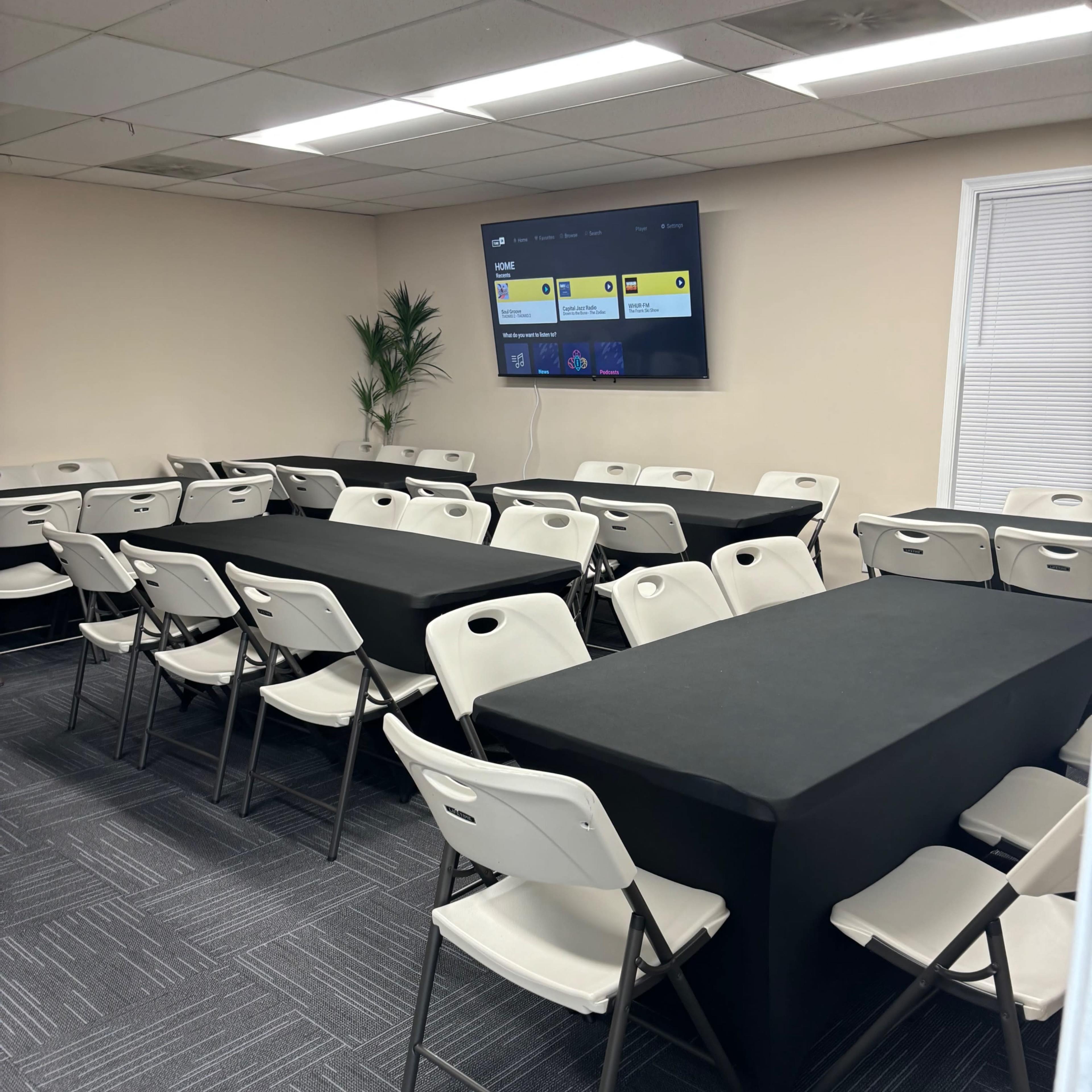 A conference room features several rows of chairs arranged around black-covered tables, with a television mounted on the wall displaying a screen interface.