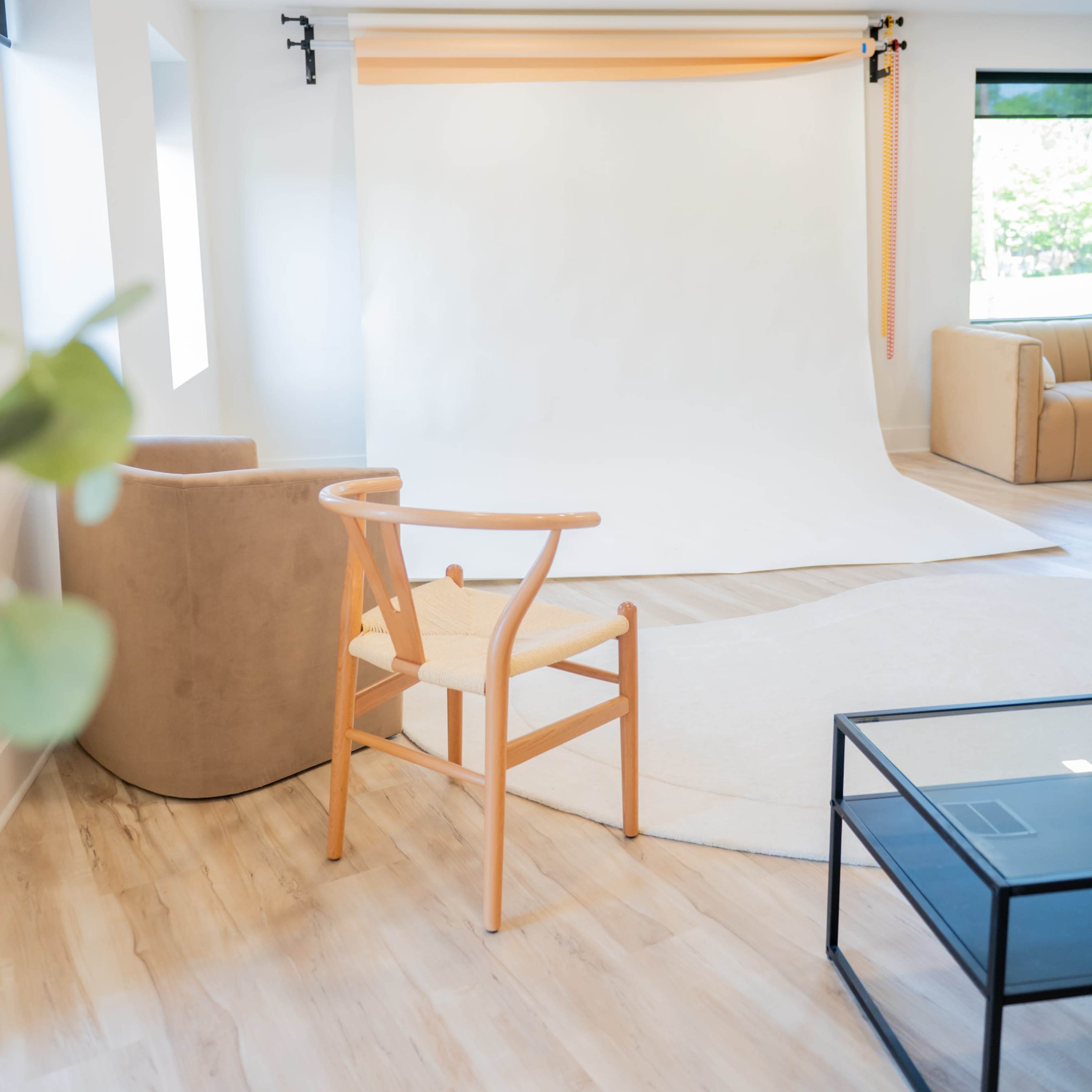 A modern photography studio with a blank backdrop, a wooden chair, a light-colored rug, and a sofa.