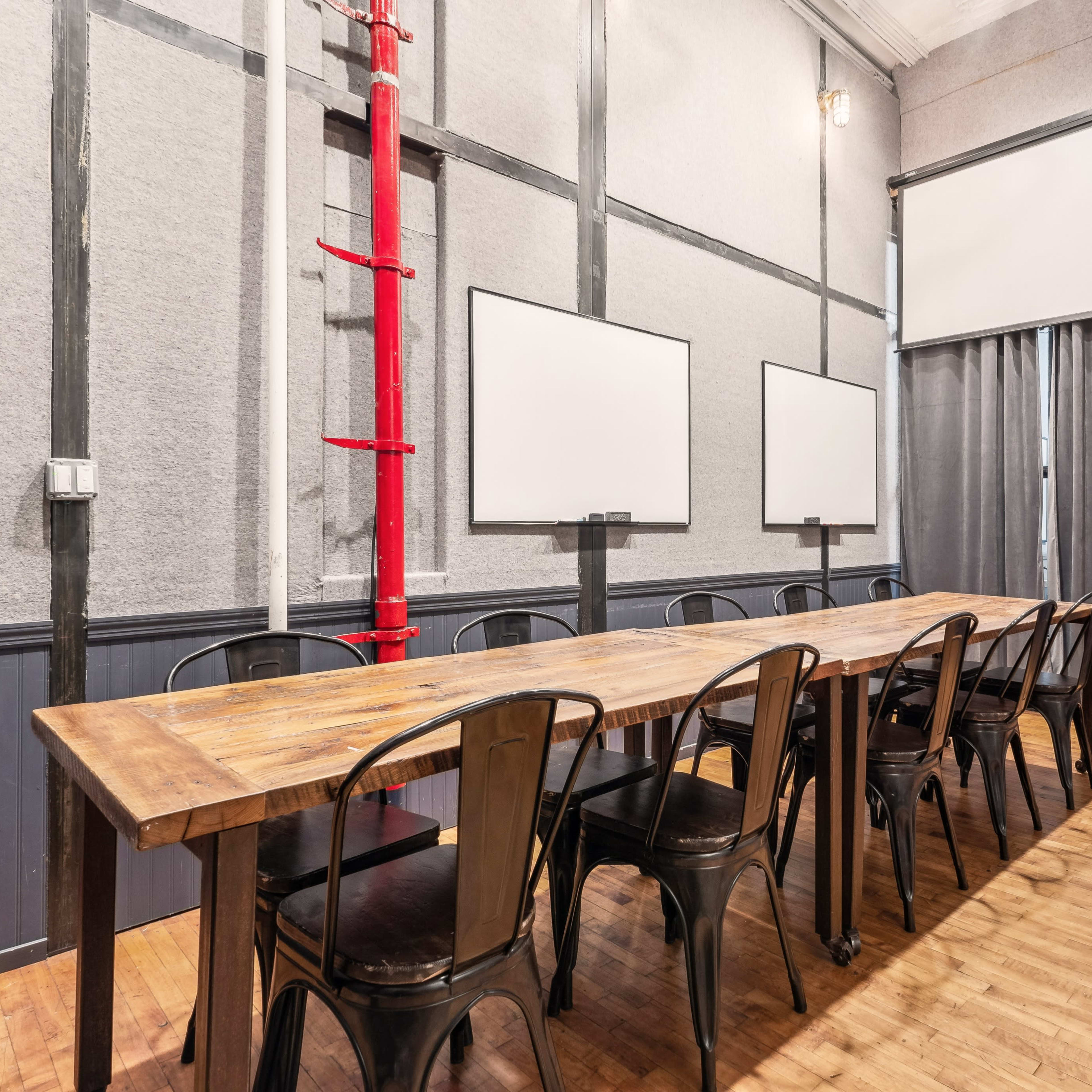 A meeting room features a long wooden table surrounded by black chairs, with gray walls and two white screens at the front.