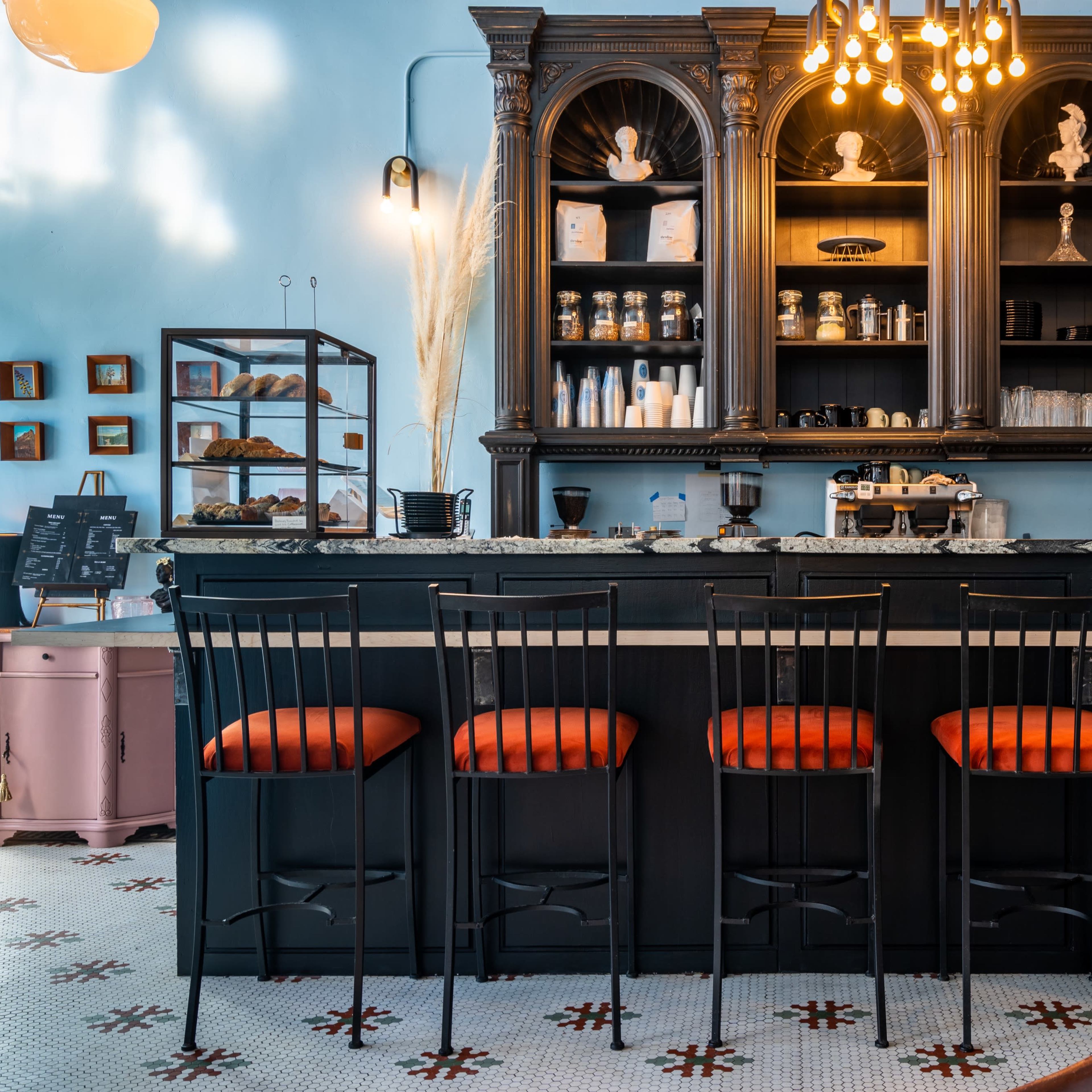 A stylish café bar features dark wood cabinetry, a marble countertop, and orange-cushioned stools lined up in front of it.