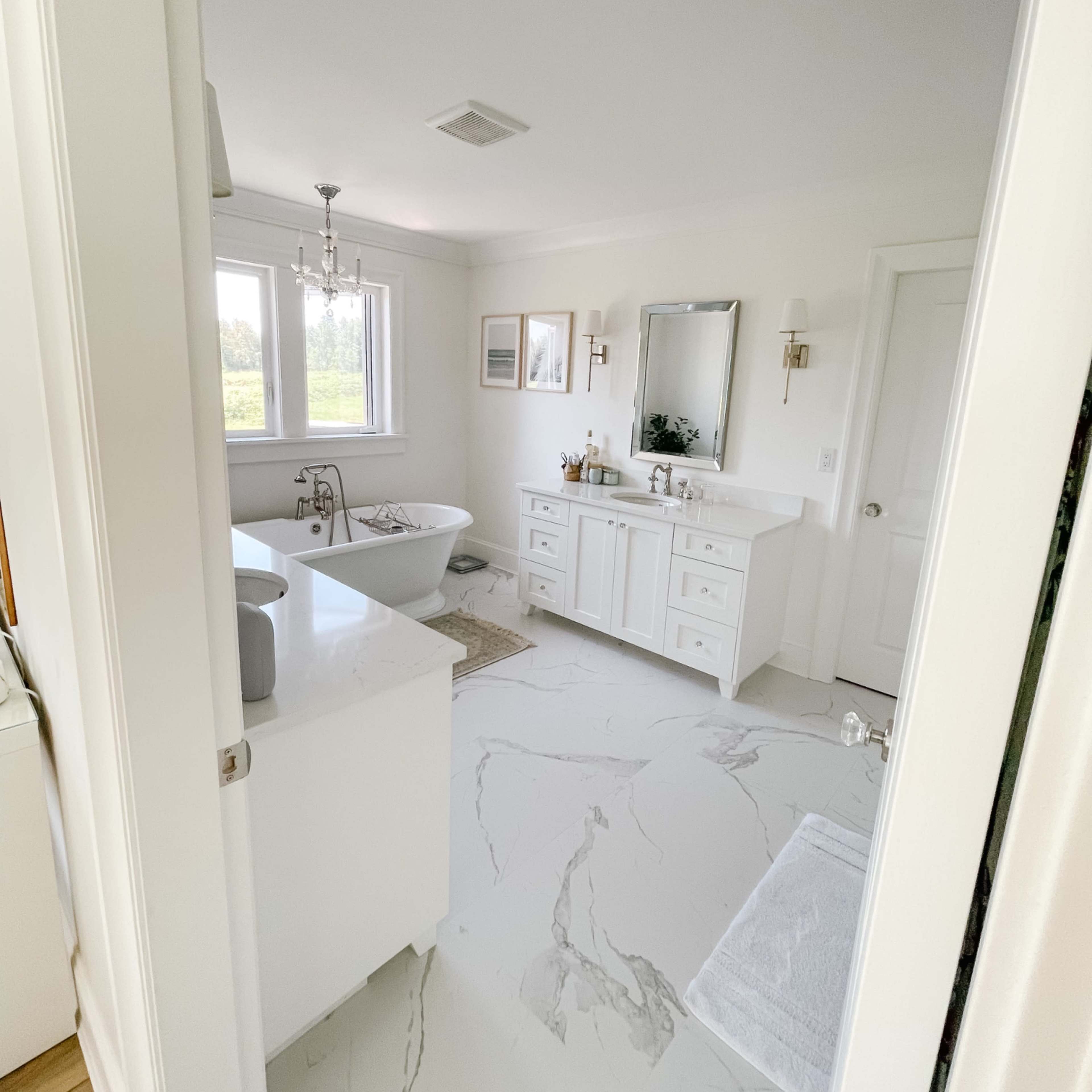 A bright bathroom features a freestanding bathtub, a double-sink vanity with a large mirror, and marble-patterned flooring.